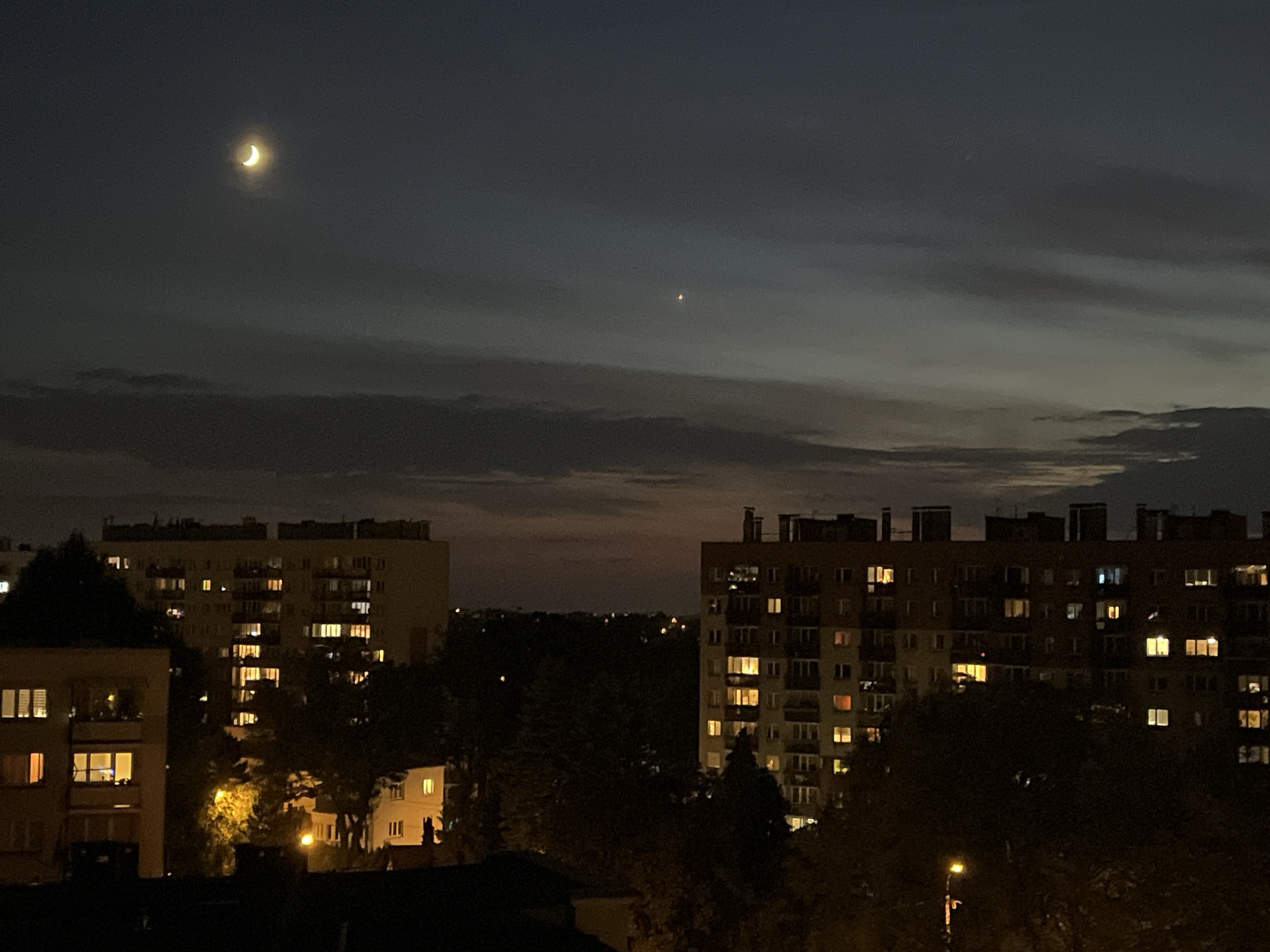A crescent moon hangs in the dark sky above a cityscape at twilight. The moon is a bright sliver of yellow in the upper left quadrant of the image. A reddish-orange point of light, likely a planet or bright star, is visible in the upper middle of the sky. Below the sky, apartment buildings are illuminated by numerous lit windows. Trees and shadows fill the foreground and obscure parts of the buildings. The buildings appear to be part of a dense urban or suburban environment. The overall mood is quiet and nocturnal.
