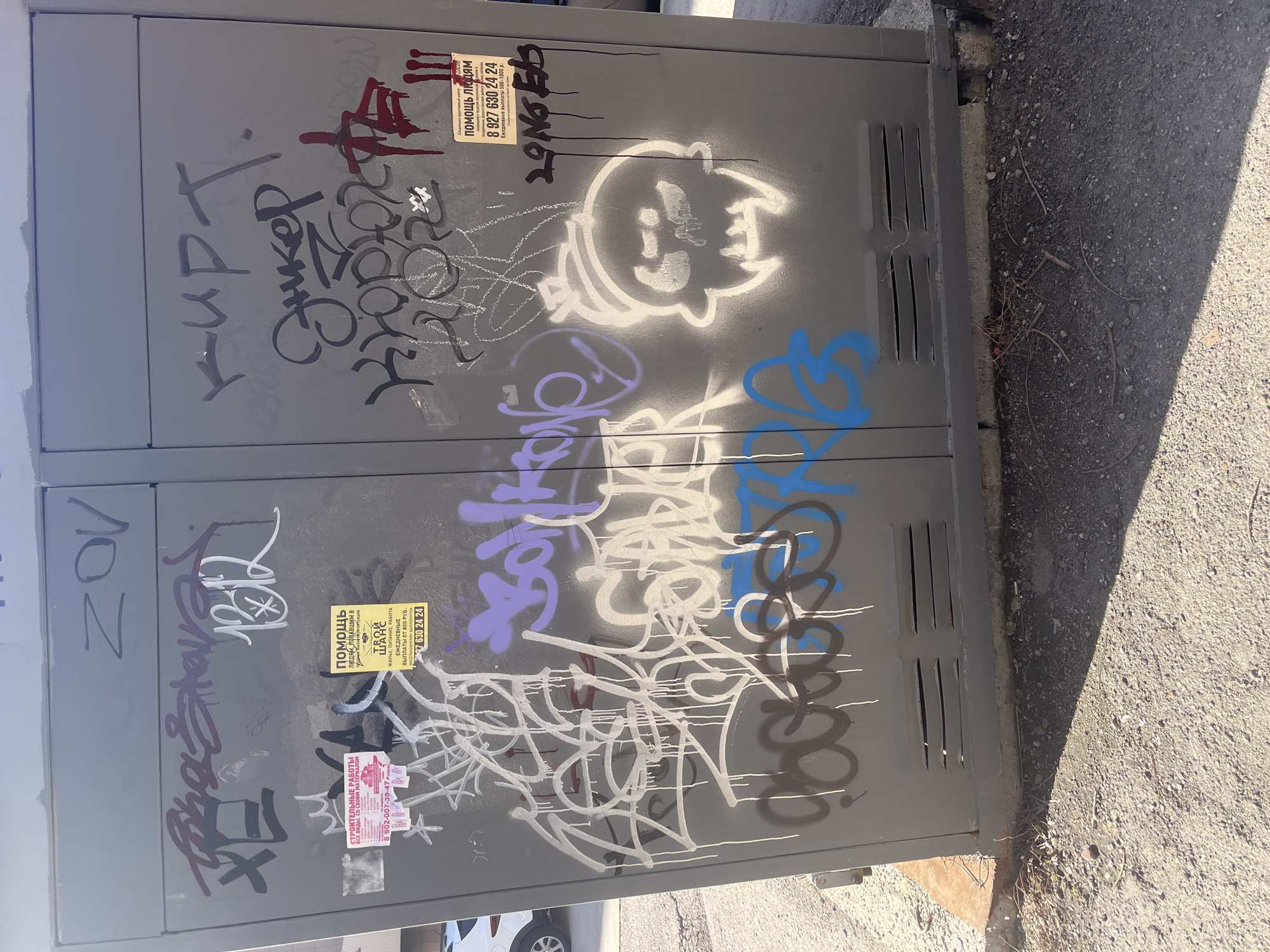 The image captures a gray metal utility box, likely an electrical or telecommunications cabinet, situated outdoors on an asphalt surface, possibly a sidewalk or roadside in Ulyanovsk, Russia. The scene is illuminated by bright daylight, casting strong shadows on the ground to the right, indicating clear and sunny weather.
The utility box is heavily covered in various forms of graffiti and several pasted advertisements. Graffiti tags are present in multiple colors, including black, white, red, purple, and blue. Notable graffiti includes "ZOV" in black on the left panel, "Энкер" (Enker) in black, and a distinct white outline of a cartoonish, skull-like or ghost-like character. Other tags are stylized and largely illegible.
Several flyers are affixed to the box:
- Two yellow flyers advertise "Помощь людям" (Help for people) and provide a phone number "8 927 630 24 24". One specifies "попавшим в трудную жизненную ситуацию" (who find themselves in a difficult life situation) and mentions "ЖИЛЬЕ, ПИТАНИЕ, РАБОТА" (Housing, food, work) with "ЕЖЕДНЕВНЫЕ ВЫПЛАТЫ ОТ 800 РУБ." (Daily payments from 800 rubles).
- A smaller pinkish flyer promotes "СТРОИТЕЛЬНЫЕ РАБОТЫ ВСЕ ВИДЫ, СО СВОИМ МАТЕРИАЛОМ" (Construction work, all types, with own materials) and lists a phone number "8 902-007-39-47".
The ground surrounding the box is coarse asphalt, with a concrete curb visible along its base. Small twigs and debris are scattered on the ground near the cabinet. The utility box itself features vertical ventilation slats on its right side. No people are visible, and the scene is static, depicting an urban fixture marked by public interaction.