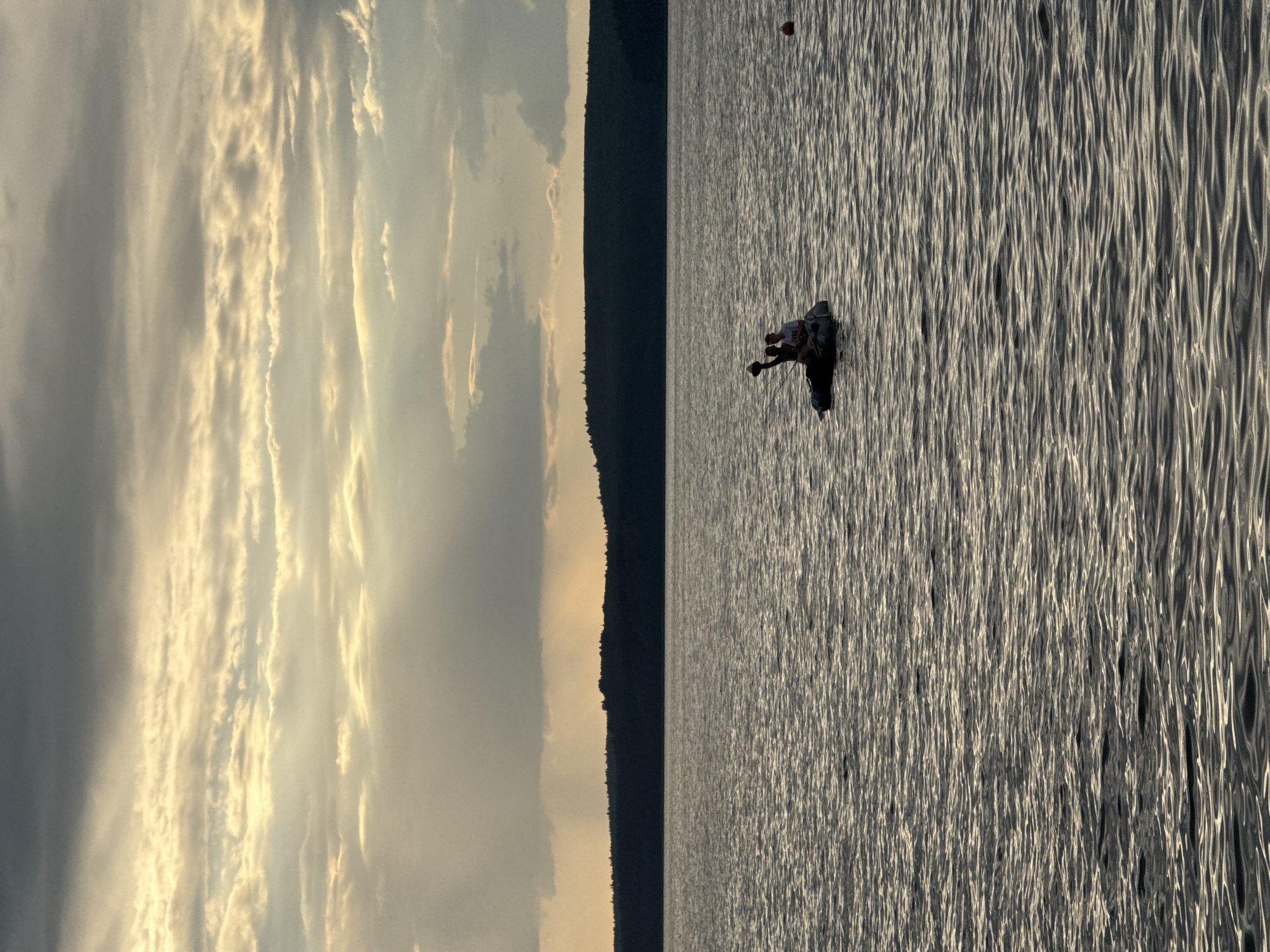 Two individuals are seated in an inflatable boat on a body of water, possibly a lake or large river. One individual is holding up an object, possibly a fishing rod or a paddle, towards the sky. The other individual is seated in front, facing forward. The boat is positioned in the middle of the frame. In the background, a dark, tree-covered shoreline stretches across the horizon, silhouetted against a dramatic sky filled with layered clouds illuminated by sunlight breaking through in rays. The water's surface is rippled, reflecting the light. A small orange buoy is visible on the right side of the frame. The scene suggests an outdoor recreational activity during either sunrise or sunset, given the lighting conditions and cloud formations. The location is identified as Votkinsk, Russia.