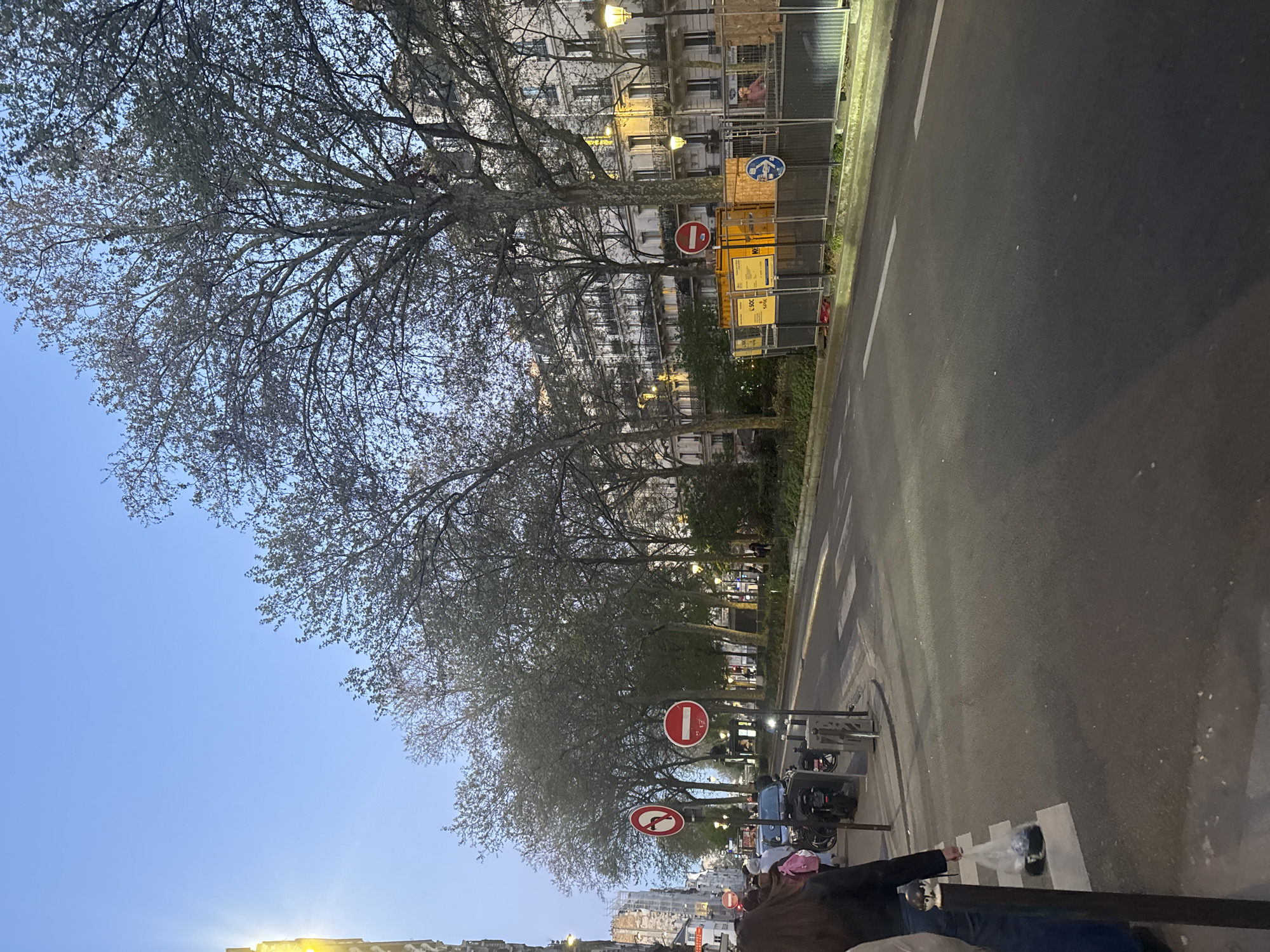 The image depicts a street scene in Paris, France, at twilight. The view is along a street lined with trees in early spring, their branches bare except for the beginnings of buds. Buildings line the opposite side of the street. There are several traffic signs visible, including "no entry" signs. Construction barriers and materials are also present. A group of people are partially visible in the foreground, standing and talking. The road is asphalt, with a white lane marking and pedestrian crossing markings. A person is carrying a bag of what appears to be trash. Streetlights are illuminated in the distance. The sky is a light blue, indicating early evening.