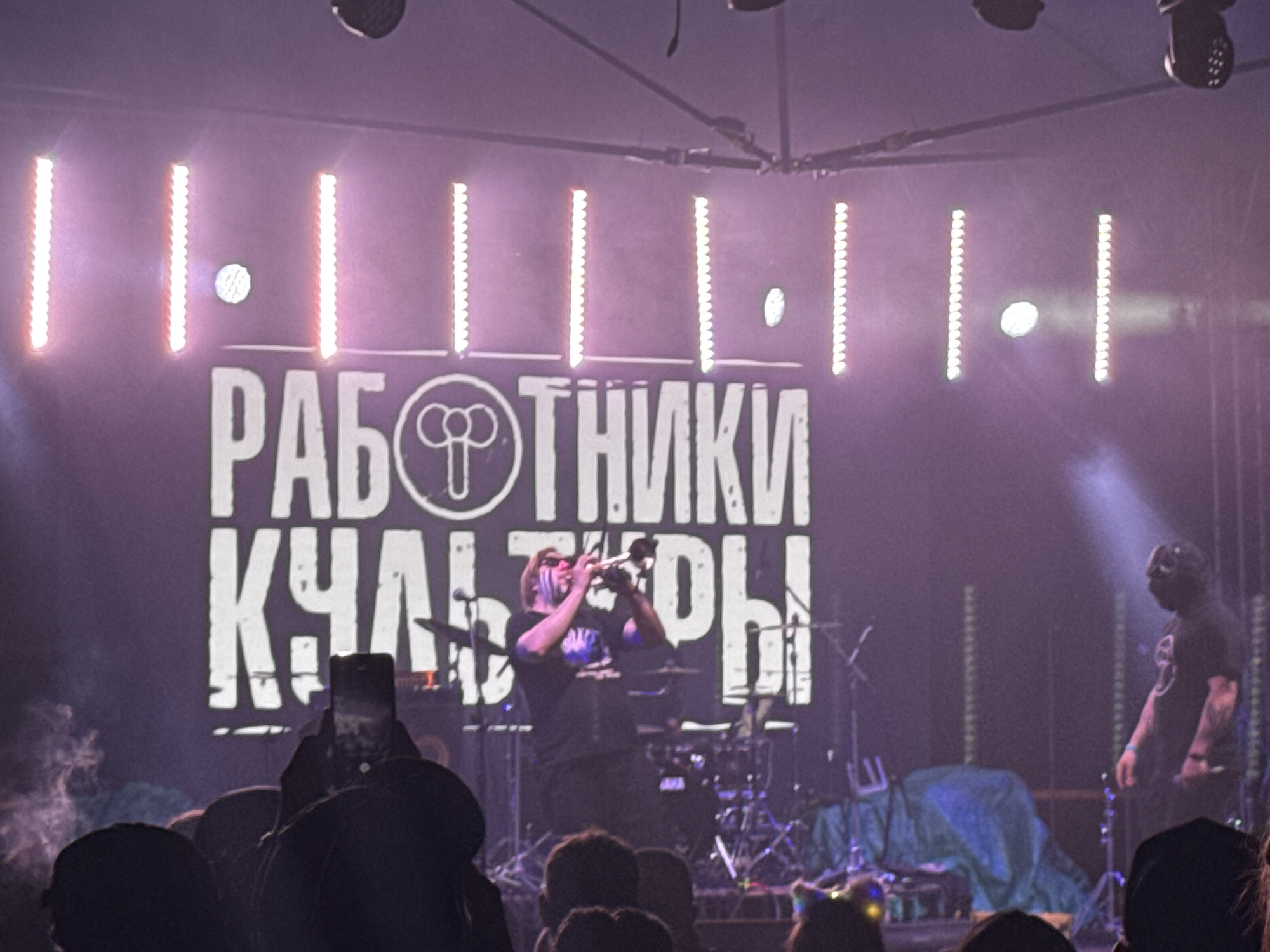This is a detailed image of a live music performance taking place on a stage in Большая Кивара, Russia. The scene is illuminated by intense stage lights, casting a predominant purple/magenta hue through what appears to be stage haze or smoke.
In the background, a large white banner or screen displays the Russian text "РАБОТНИКИ КУЛЬТУРЫ" (Workers of Culture). The letter "O" in "РАБОТНИКИ" is replaced by a stylized circular logo resembling three interconnected loops or a bow tie. Above and beside the text, multiple vertical LED light strips, some actively illuminated, and several circular stage lights are visible.
On the stage, there are at least two male performers.
1. **Central Performer:** Positioned slightly left of center, a male individual with light-colored hair and distinctive white or silver face paint (streaks across his eyes and cheeks) is actively playing a brass instrument, likely a trumpet, held to his mouth with both hands. He is wearing a dark t-shirt with a faint design and dark pants. A drum set, including cymbals and drum shells, is partially visible behind him. A microphone stand is also nearby.
2. **Right Performer:** To the right of the central performer, another male individual stands. He is wearing a dark t-shirt, dark pants, and a dark mask that completely covers his face. Tattoos are visible on his right forearm. He is standing relatively still, facing slightly towards the left, with his hands down at his sides.
Other stage elements include a blue or greenish tarp draped over some equipment on the right side of the stage and various stands and cables.
In the immediate foreground, the heads and shoulders of numerous audience members are visible, indicating a crowded event. One audience member, slightly left of center, is holding up a black smartphone, actively recording or photographing the performance, with the phone screen displaying an image. Some audience members on the right side of the frame have glowing accessories, such as light-up headbands or ears. The atmosphere is hazy, with visible light beams from the stage lighting.