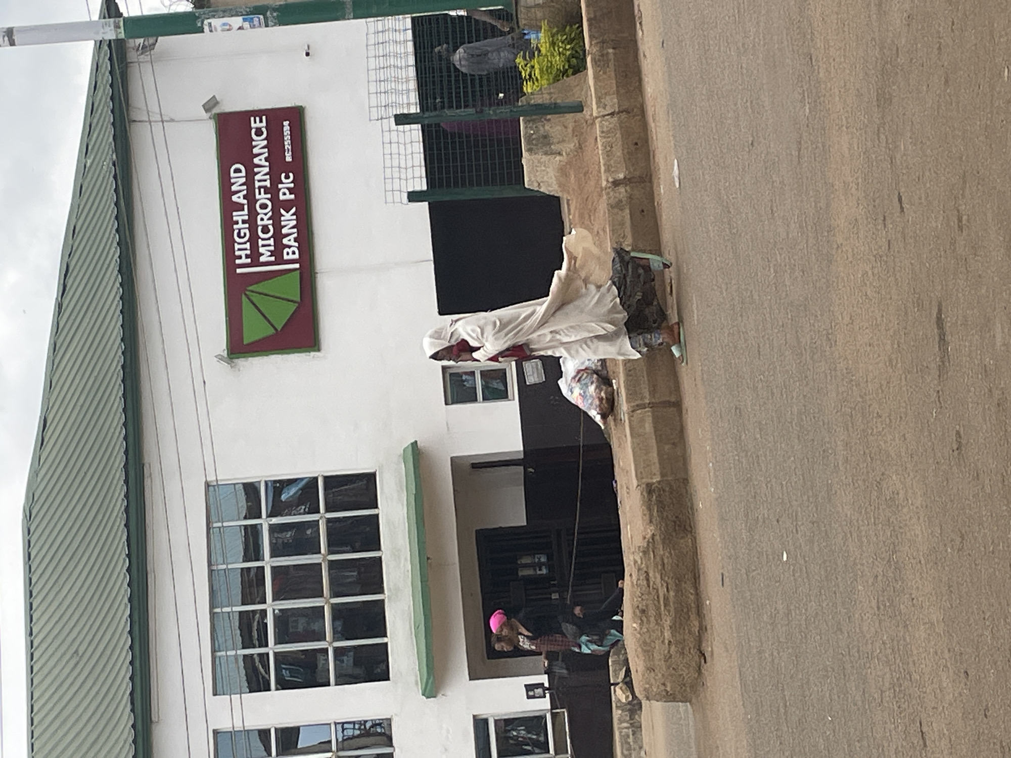 The image shows a street scene in Bukuru, Nigeria, outside Highland Microfinance Bank Plc. A building with a green roof and white facade houses the bank. Large, multi-paned windows are visible on the building, reflecting the surroundings. A prominent sign for "HIGHLAND MICROFINANCE BANK Plc" is mounted on the wall.
Several people are present in the scene. In the foreground, a person wearing a light-colored, flowing garment and a head covering walks across the sidewalk, carrying a red item. To their left, two individuals are walking near the entrance of the building. One is wearing a dark top and a pink head covering, and the other is wearing a patterned shirt. Further in the background, through a metal fence, other figures are partially visible.
The environment includes a paved street, a concrete curb, and a raised, rough-hewn concrete barrier. To the right of the bank, there is a small patch of green vegetation. Trash bags are visible on the sidewalk near the walking individuals. The overall lighting suggests an overcast day.