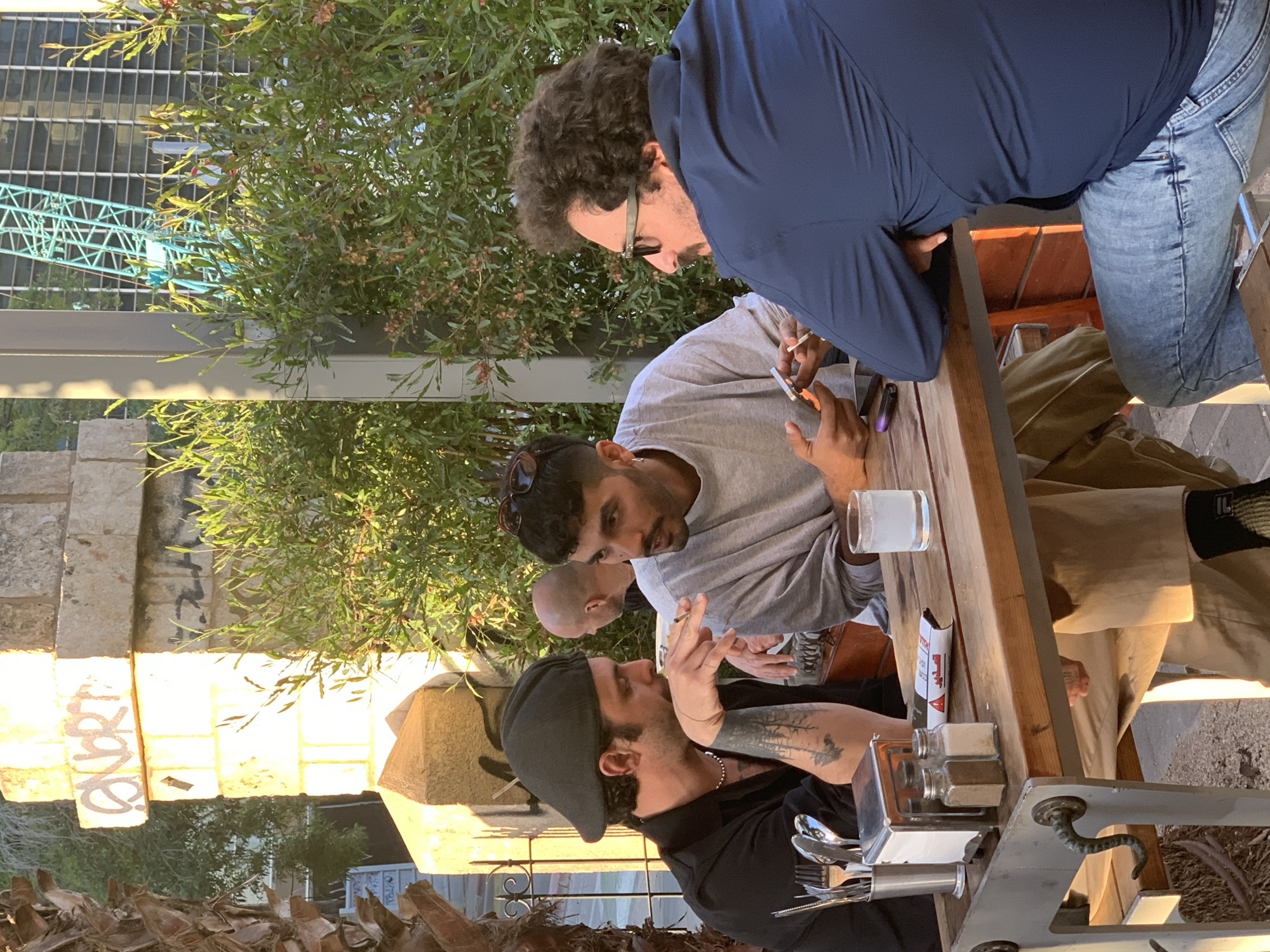 Four men are seated at an outdoor wooden table, engaged in conversation. One man is rolling a cigarette. The table is set with cutlery, a salt and pepper shaker, a glass of water, and a pack of cigarettes. The men are outdoors, with trees and a building visible in the background. A crane can be seen in the background. Based on the architecture and the presence of a palm tree, this location is most likely in Tel-Aviv, Israel. Graffiti is visible on a nearby wall.