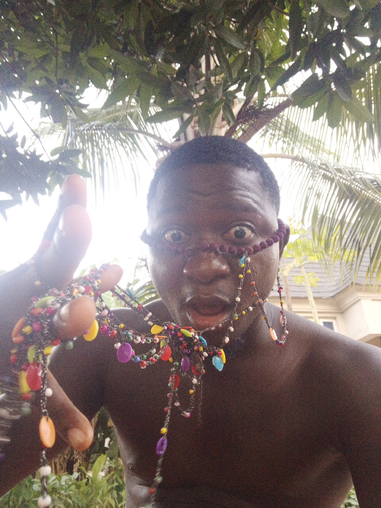 A close-up shot shows a Black man holding a multi-colored beaded necklace, some strands draped across his face and head. The background features lush green trees and foliage with glimpses of a building. The image was taken in Akwanga, Nigeria.