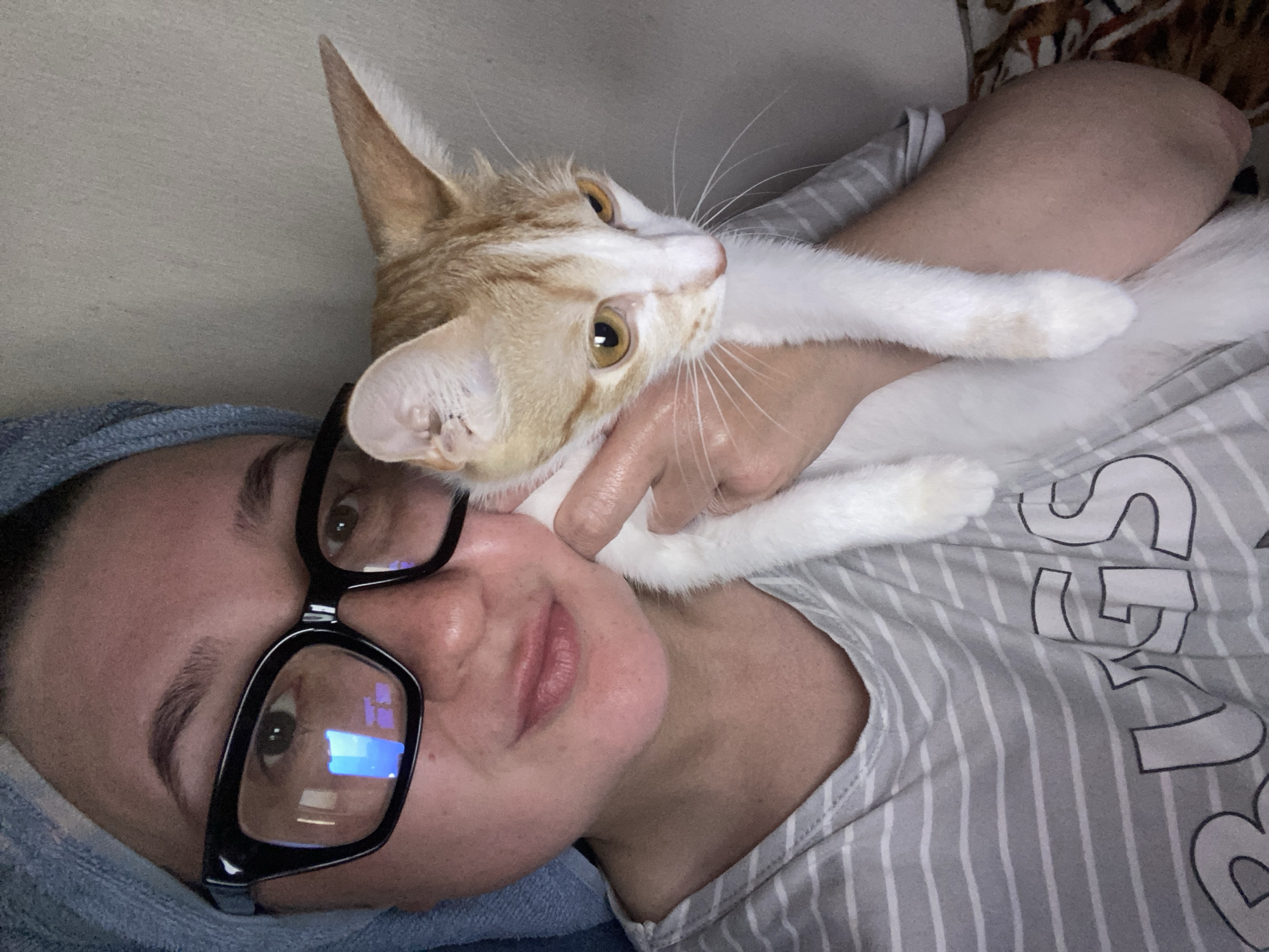 The image shows a close-up selfie of a person holding a cat. The person is wearing glasses and a towel wrapped around their head, suggesting they may have recently showered. The cat is an orange and white tabby. The background is a plain wall. The image does not contain any specific information to determine the location within the city of Chaikovsky, Russia.