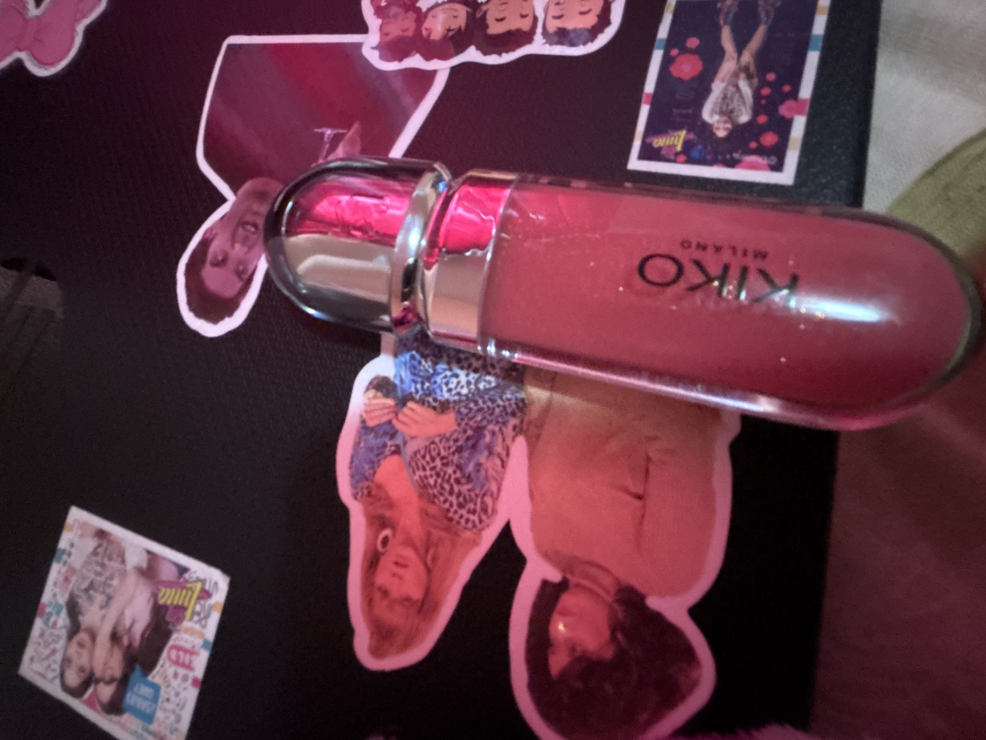 A close-up, medium-shot image shows a black surface adorned with various stickers and a tube of Kiko Milano lip gloss. The lip gloss is positioned horizontally in the center of the frame, its pink color and metallic cap prominent. The stickers include images of celebrities, a group of people, and a sticker with the word "Luna" and other text and graphics in the lower left. The environment is undefined. The image does not contain any information about Garmen, Bulgaria.