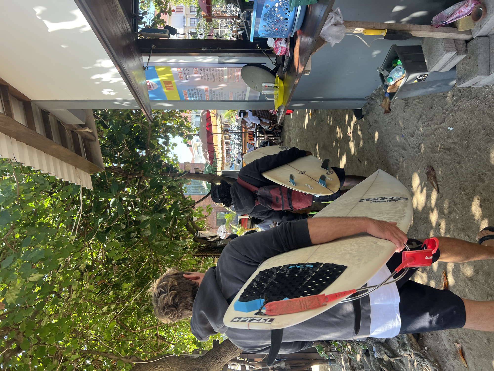 In Kuta, Indonesia, a scene unfolds on a sandy beach path adjacent to a building with an open-air serving counter. Two individuals are walking away from the camera, carrying surfboards. The person in the foreground, viewed from behind, wears a dark grey hoodie and black shorts, with a lighter t-shirt visible underneath. They are carrying a light-colored surfboard with a red leash and fins. Behind them, another person, also seen from the back, wears a dark hoodie and a red backpack, and is carrying a similar light-colored surfboard.
The building appears to be a small cafe or food stall, with a menu board displaying prices for coffee, tea, and food items like fried rice and noodles. Various items are visible on the counter, including a yellow plate with a glass, a blue plastic basket, and some produce. In the background, more beachgoers can be seen under umbrellas, and rows of parked motorcycles suggest a bustling area. Lush green trees with dappled sunlight overhead provide shade. The overall environment suggests a typical day at a popular beach destination.