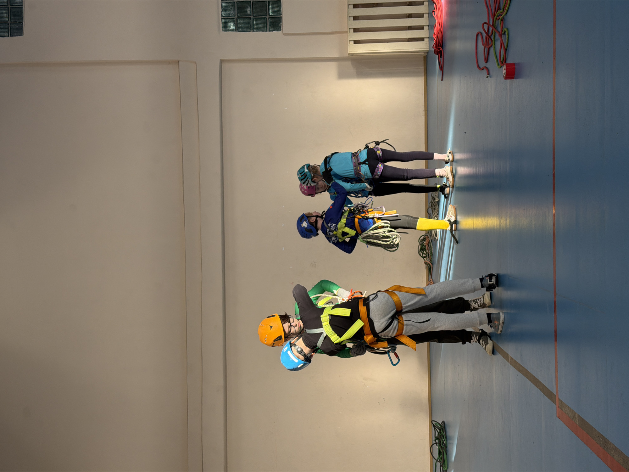 Three individuals are standing on a blue floor in a large room with light beige walls. The room appears to be an indoor climbing or training facility, given the presence of climbing ropes and gear. Two ventilation window blocks are visible near the top of the left wall, and a wooden slatted structure is on the right wall.
The three individuals are wearing climbing harnesses and helmets. The first individual, closest to the viewer, wears a blue helmet, a green top, a grey climbing harness with yellow straps, and grey pants. The second individual has an orange helmet and black top. The other two individuals are wearing blue climbing attire with green and pink helmets, respectively.
The individuals appear to be preparing for or practicing a climbing activity, as evidenced by the ropes and harnesses. The floor is marked with a red or orange stripe. Various climbing ropes are hanging or lying around the room.
Based on the available information, the location appears to be an indoor training facility that could be in or near the city of Berezniki, Russia, if this is the location of the image.