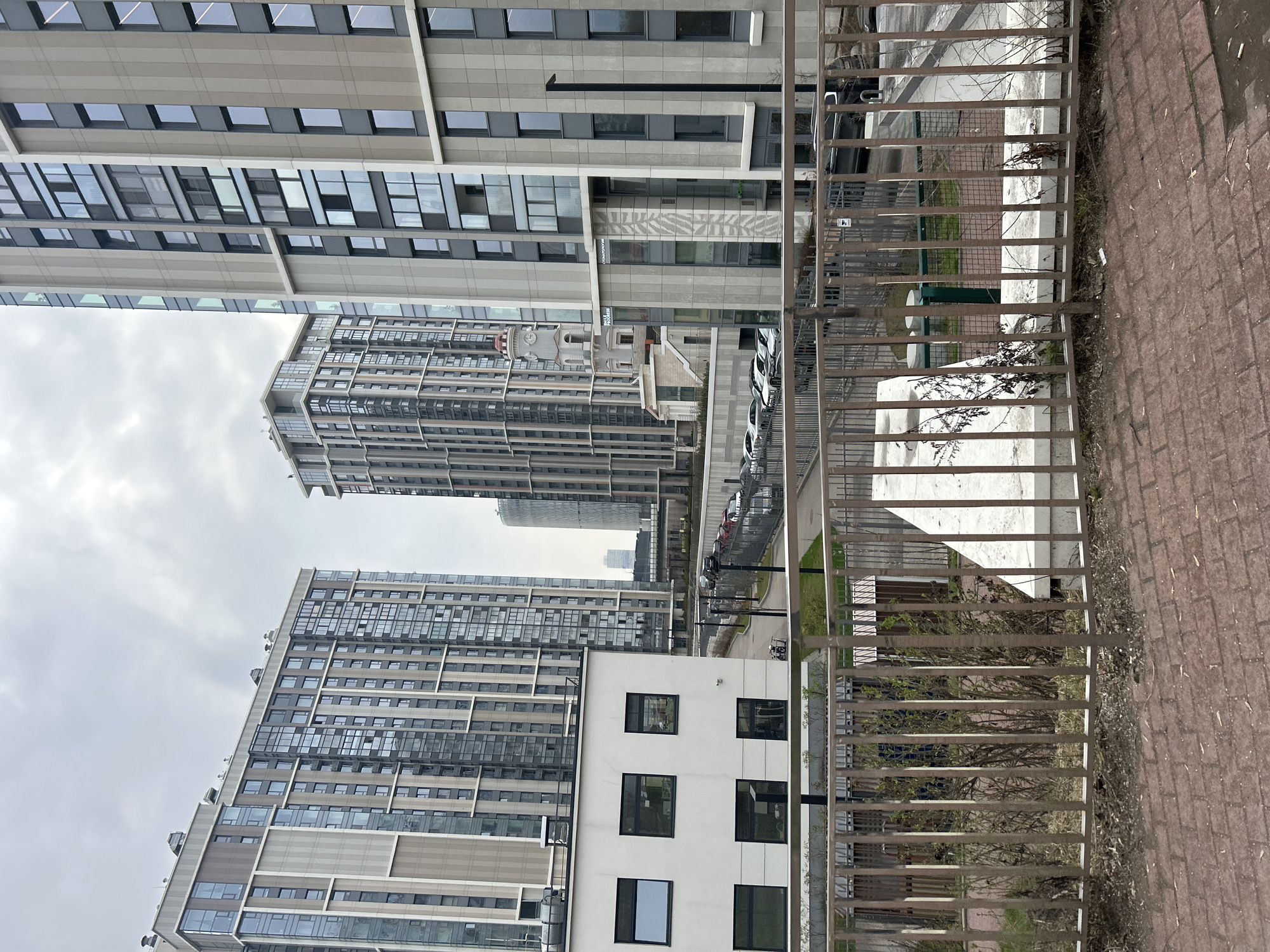 The image depicts a modern urban landscape in Moscow, Russia, under an overcast sky. Several tall, contemporary buildings with multi-story glass windows and facades of varying shades of grey and white dominate the scene. Their architecture is sleek and geometric.
In the foreground, a metal fence runs horizontally, separating a red brick-paved area from a lower section that includes a strip of sparse, bare vegetation and some small, immature trees. Beyond this, a road or parking area features several parked cars, mostly light-colored. A smaller, white building with square dark windows is visible to the right, adjacent to the fence line.
Among the prominent structures, one building displays the text "МЕДИЦИНСКИЙ ЦЕНТР" (Medical Center) and "CLINIC" on its facade, indicating its purpose. This building also features a distinct geometric pattern integrated into its design. Further in the background, another building with a small dome and a clock tower adds a unique architectural detail to the modern skyline.
The time of day is daytime, though the consistently grey sky suggests cloudy or overcast weather. No people are explicitly visible, but the presence of parked cars implies daily activity and habitation within this developed city environment. The scene portrays a functional and aesthetically modern district.