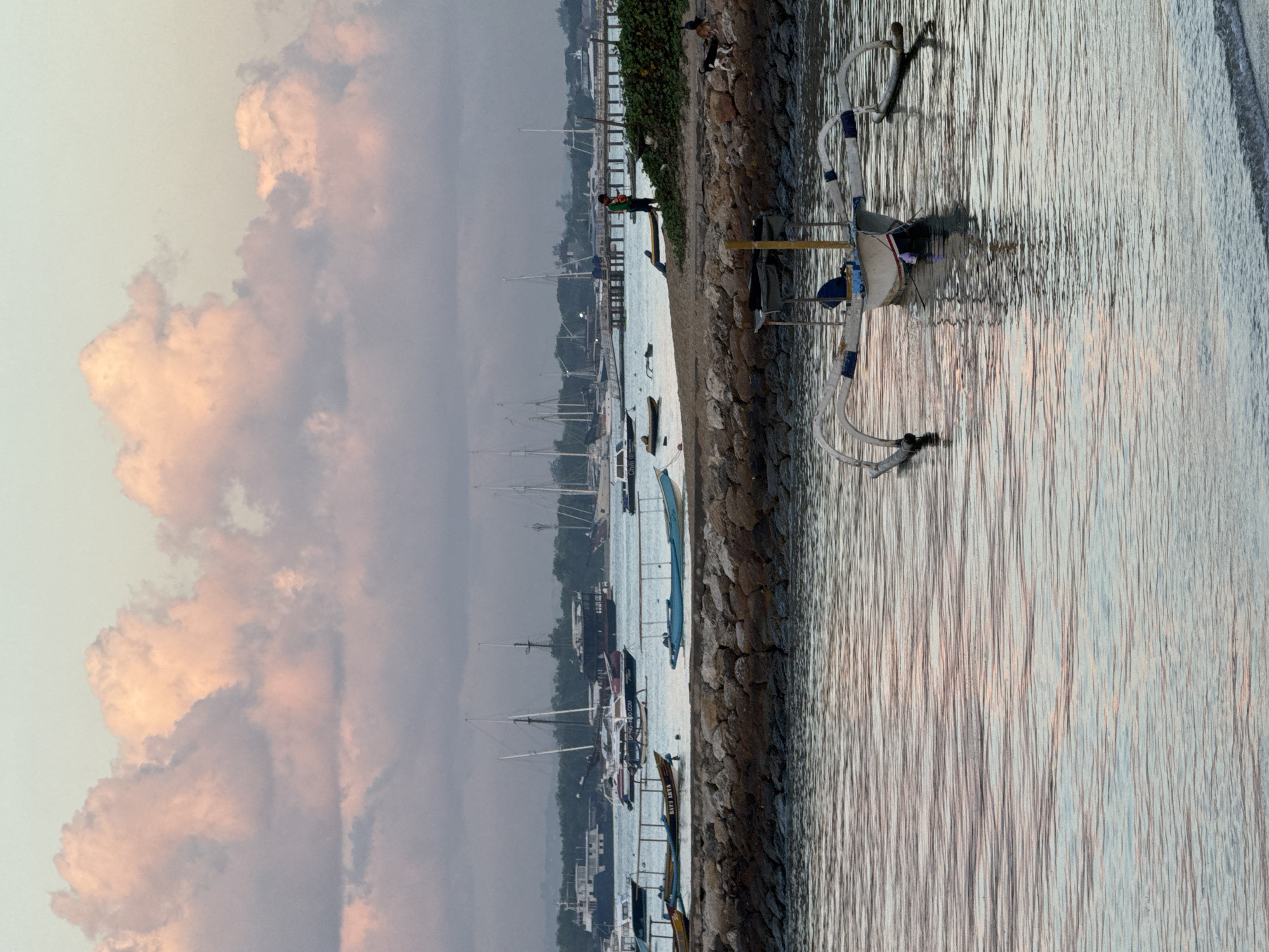 The image depicts a serene harbor scene in Denpasar, Indonesia, likely during sunrise or sunset given the soft, pastel hues of the sky and clouds. In the foreground, a traditional Balinese outrigger boat, known as a jukung, is moored in calm, rippling water. The boat is white with blue and red trim and has characteristic outrigger floats.
Beyond the jukung, a stone breakwater separates the foreground water from a sandy shore. On the shore, several other boats are visible, including smaller fishing boats and larger vessels further out in the harbor. Some of these boats are sailboats, with tall masts reaching towards the sky.
In the middle ground, a person in a green shirt is standing on the shore, near a line of trees and a pier extending into the water. Two dogs are also present on the shore, one appearing to be a black and white mixed breed and the other a darker canine. The background shows a hazy treeline and a sky filled with soft, illuminated clouds, tinged with pink and orange, suggesting a peaceful end to the day or the gentle start of a new one. The overall atmosphere is tranquil and captures the essence of coastal life in Denpasar.