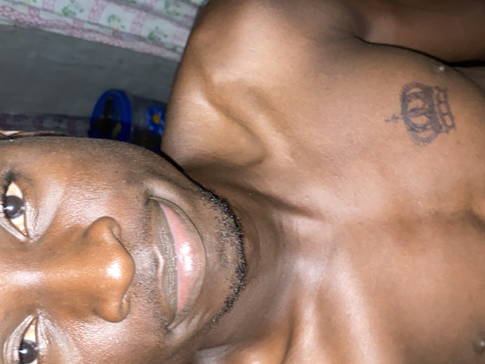 A close-up, angled image shows a dark-skinned person lying down. Part of the person's face, including eyes, nose, mouth and facial hair are visible in the lower-left portion of the image. The upper-right portion shows their upper chest and shoulder area. A tattoo is visible on the upper chest. The background contains bed linens, including what appears to be a bed sheet, and a blue and yellow container. There is no location in Barden Yana, Nigeria evident in this image.