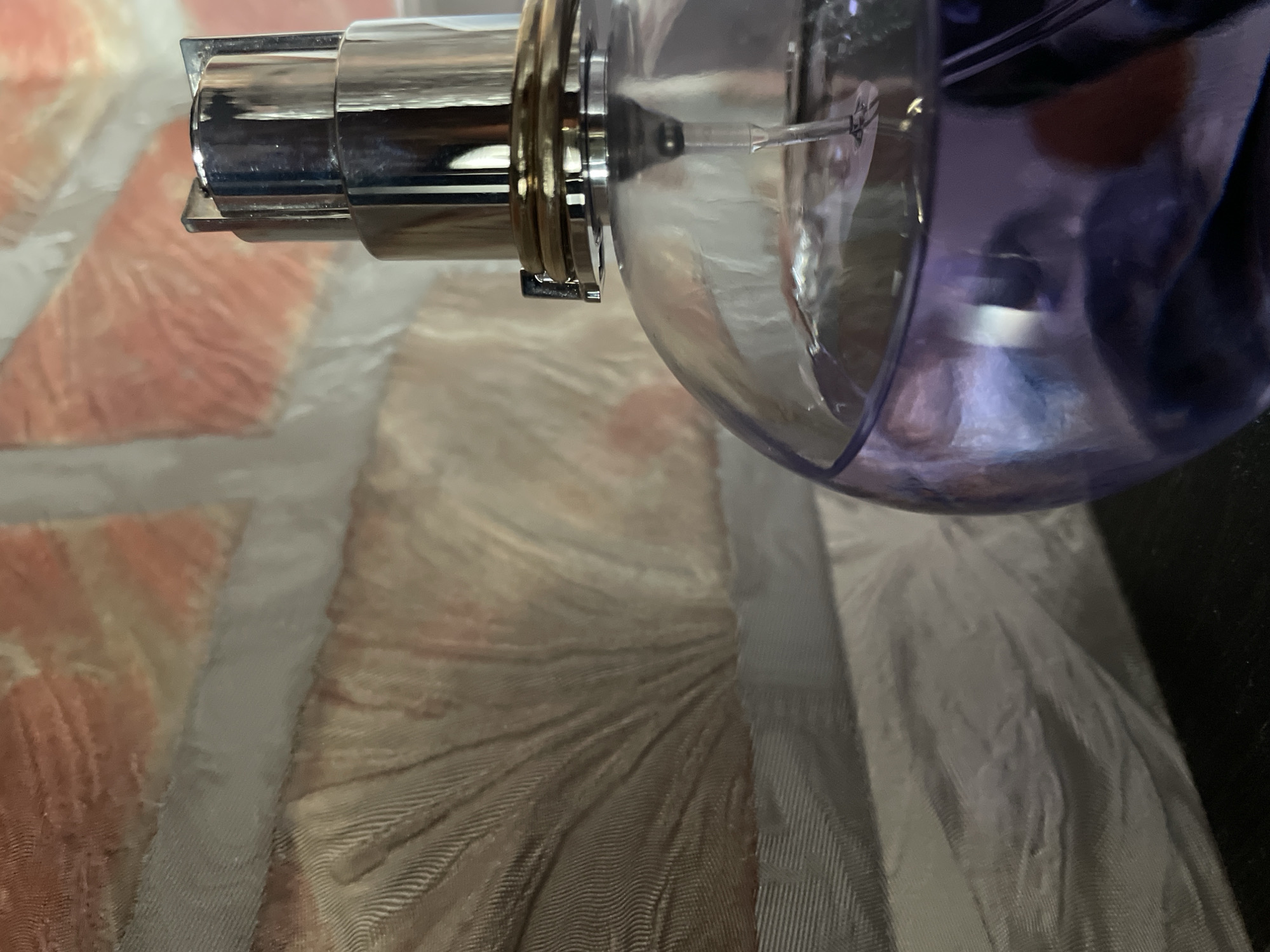 The image shows a close-up of a perfume bottle with a metallic pump mechanism and a transparent purple-tinted bulb. The bottle is positioned against a textured fabric surface that appears to be a mix of light-colored patterns, possibly on a bedspread or cushion. The pump is on the left side of the frame. The environment is indoors. There is no indication of the location in Izhevsk, Russia.