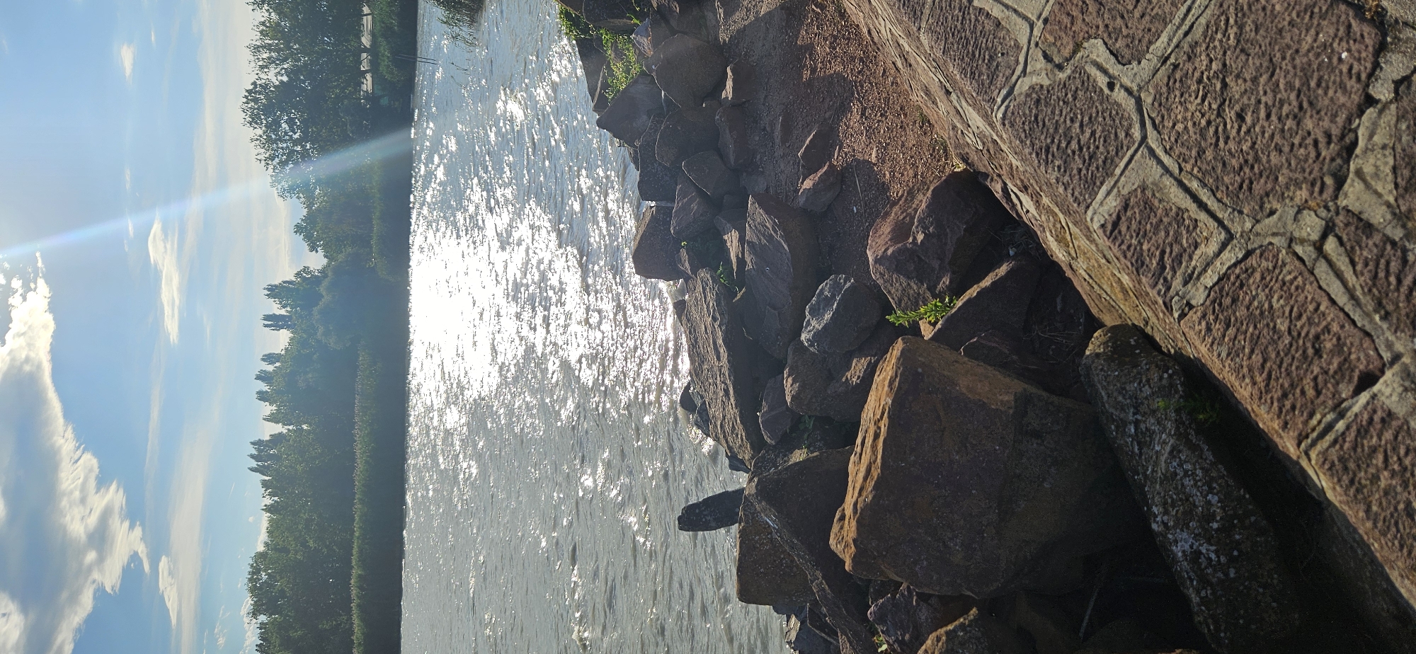 The image depicts a sun-drenched waterfront scene with a rocky embankment and a calm body of water. The sky is a vibrant blue with scattered white clouds, and the sun's rays create a sparkling effect on the water's surface. The embankment is covered with large, rough stones, some of which are reddish-brown and others are darker gray. Green vegetation, including small plants and grasses, grows between the rocks. In the foreground, a paved walkway with patterned stones is visible on the right side. The opposite shore features a dense line of trees and vegetation. The image is taken from a low angle, emphasizing the texture of the rocks and the expanse of the water. There are no people or animals visible in the frame. The setting suggests a natural or semi-natural waterfront environment, possibly a park or a recreational area. Balatongyörök is a village located on the northern shore of Lake Balaton in Hungary, known for its scenic beauty and waterfront access, which aligns with the visual elements present in the photograph.