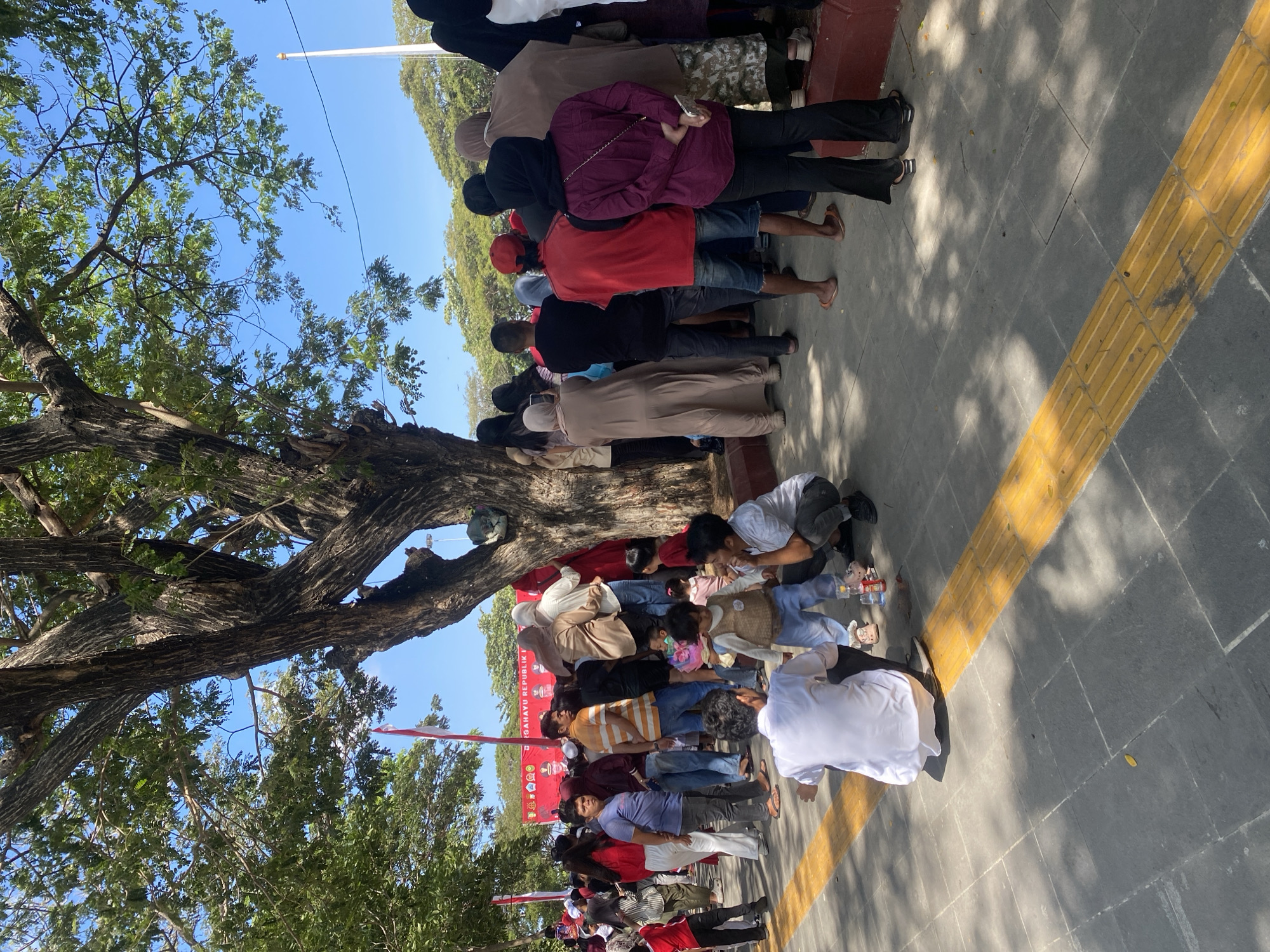 The image depicts an outdoor scene in the city of Bantaeng, Indonesia, likely a public square or park, given the presence of large trees and a paved walkway. A crowd of people, dressed in a variety of casual clothing, are gathered. Many are wearing head coverings, suggesting cultural or religious observance. Some individuals are standing, while others are sitting or kneeling on the ground. Children are also visible, some interacting with adults. In the background, a red banner with white text and graphics is displayed, possibly indicating an event or celebration. The environment features mature trees with lush green foliage, casting shadows on the ground. The sky is clear and blue. The overall atmosphere appears to be one of a public gathering or event.