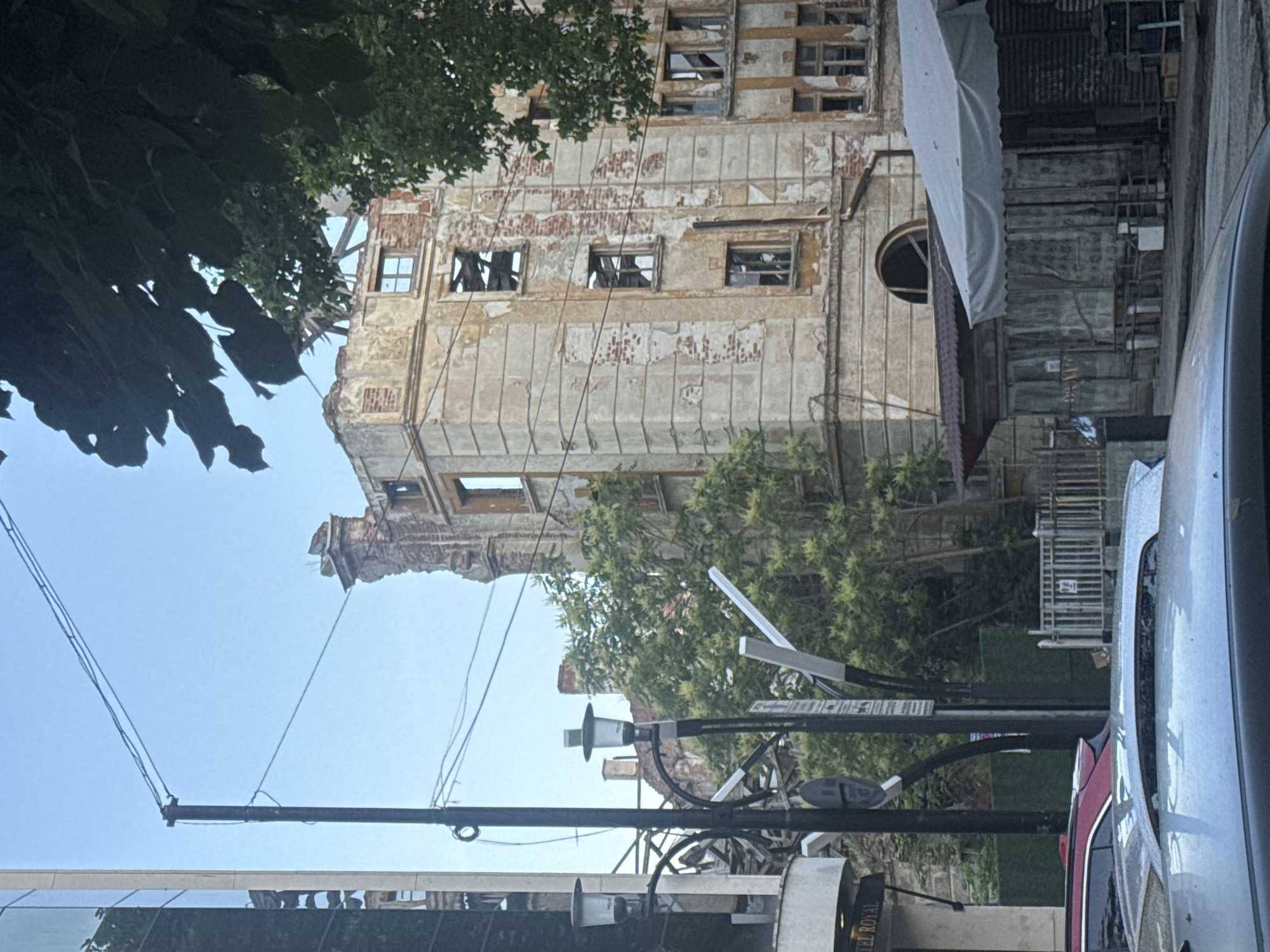 The image captures a severely dilapidated, multi-story building from a street-level perspective, viewed under a clear, bright blue sky. The building, likely an old hotel or commercial structure, shows extensive damage with large sections of its plaster facade crumbling away, revealing the underlying brickwork. Many windows are broken, appearing as dark, empty frames. A significant portion of the building's upper left side is heavily ruined, resembling a partial collapse.
In the immediate foreground, a robust black utility pole stands on the right, adorned with multiple wires, streetlights, and signs. A silver car is partially visible on the far right, with a glimpse of a red vehicle behind it. To the right of the main building, a white tarp or awning covers an area, with various wooden and metal structures visible beneath and behind it. Lush green trees frame the upper left corner of the image and also appear in the mid-ground, contrasting with the worn architecture.
The scene takes place during daylight hours, suggested by the bright sky. No specific human activity is depicted. A faint "HOTEL ROYAL" text is visible on a vertical sign attached to the utility pole. The overall impression is that of an older urban environment, possibly in a state of neglect or awaiting renovation, in a location like Plovdiv, Bulgaria.