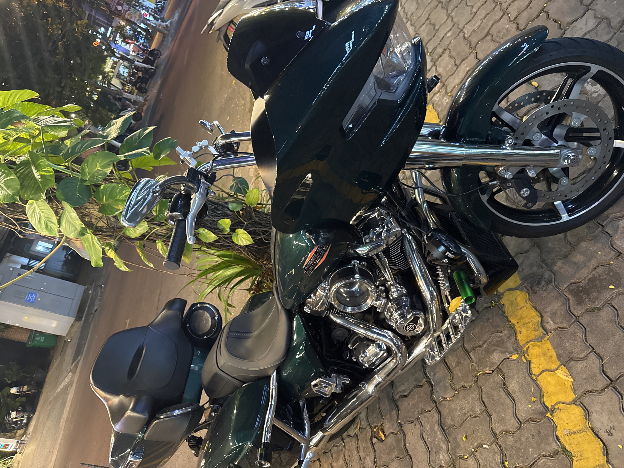 A parked, custom Harley-Davidson motorcycle dominates the foreground. The bike is dark green, with extensive chrome detailing, including the engine components, exhaust, and front forks. The motorcycle has a large front fairing, a two-piece seat, and aftermarket mirrors. A green potted plant obscures part of the motorcycle's handlebars. The motorcycle is parked on a paved brick surface with yellow painted markings. In the background, blurred street scenes suggest an urban environment, likely a city in Vietnam. Blurred storefronts and traffic lights are visible in the distance. The presence of the potted plant and the lighting indicate that the scene is outdoors and at night. Based on similar images found online, the location is likely in Đà Nẵng, Vietnam.