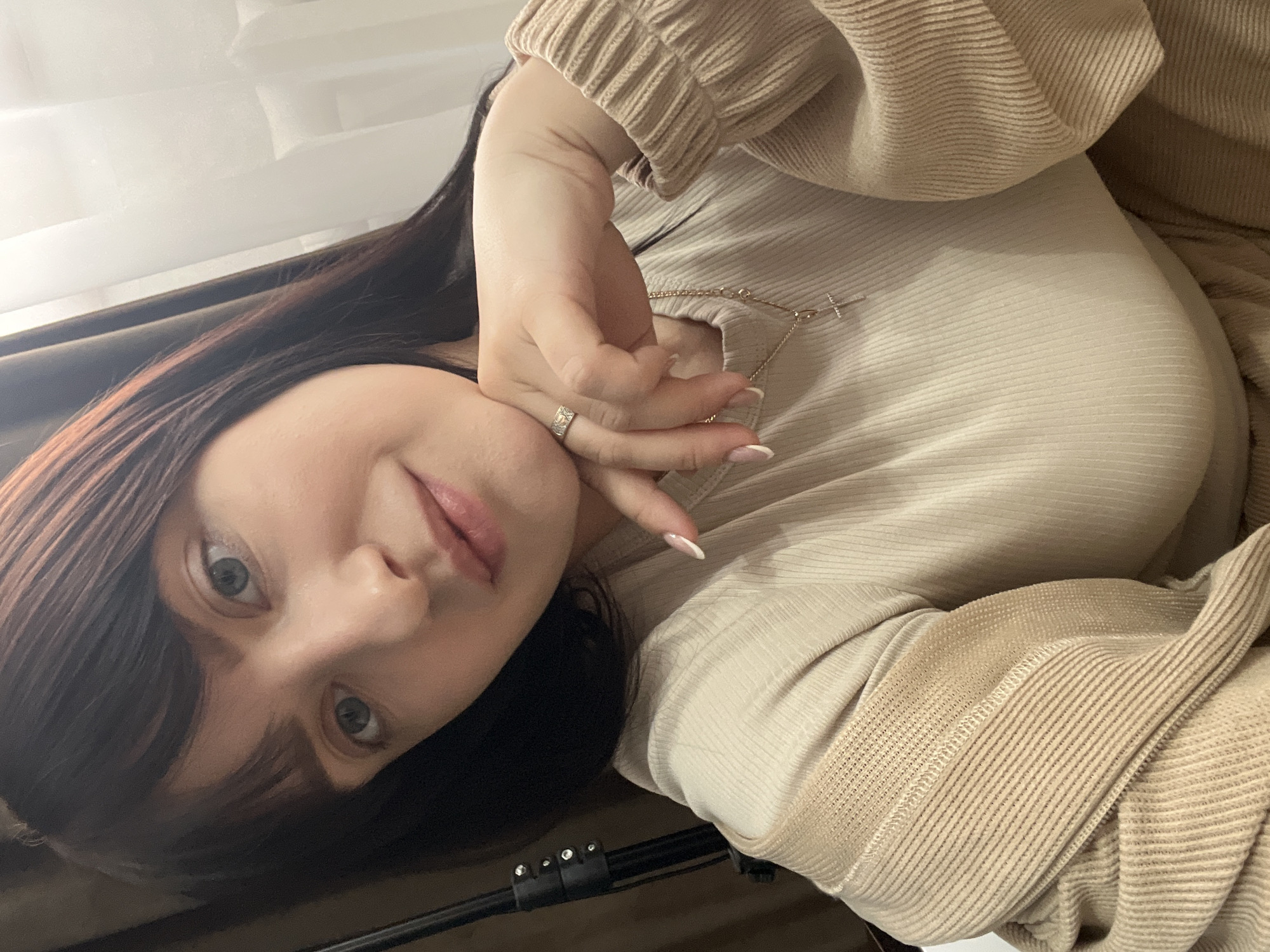 A woman with fair skin, dark hair with bangs, and light-colored eyes is looking at the camera. She is wearing a light beige ribbed shirt or dress. Her left hand is near her face, with a ring visible on her ring finger. A thin silver necklace with a cross pendant is around her neck. Part of a dark brown curtain is visible in the background, along with a white or light-colored wall. A black object is in the bottom of the frame. The image appears to be taken indoors. There is no indication of the city of Марковский, Russia.
