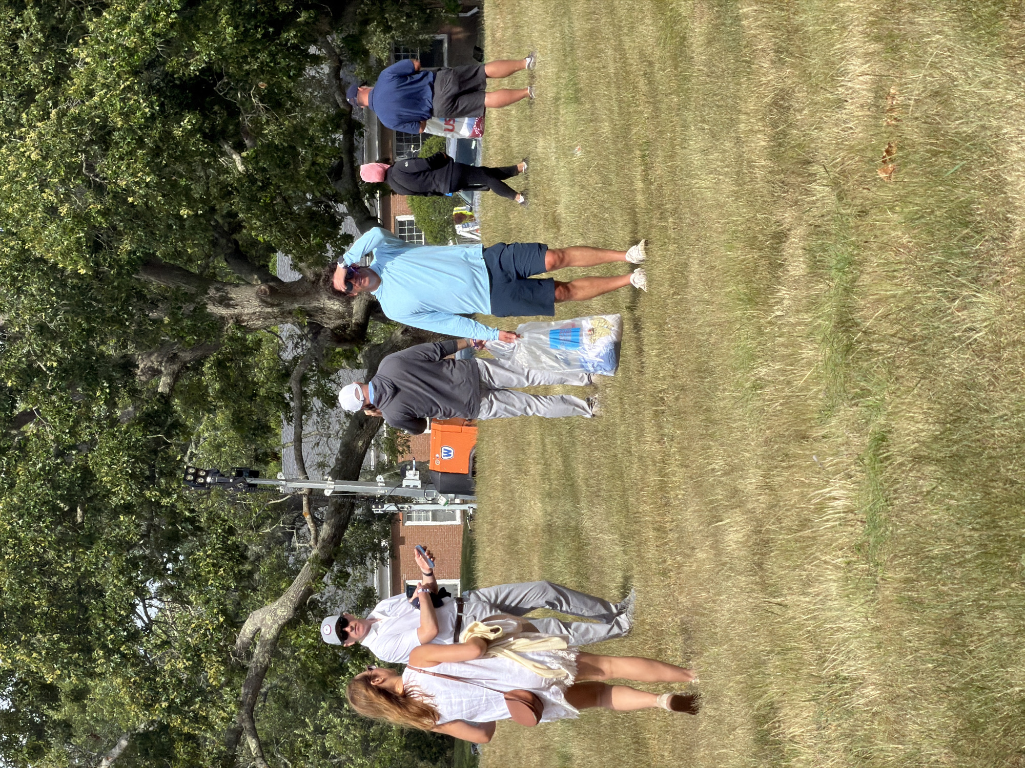 In the Town of Southampton, United States of America, a group of people are gathered on a grassy field. The image captures a sunny day with a large tree providing shade.
In the foreground, a woman in a white dress with a brown purse and a light-colored scarf is walking away from the camera. Beside her, a man in a white polo shirt and gray pants is holding a phone in his left hand and a dark object in his right. He is wearing sunglasses and a white cap.
Further back, another man in a gray hoodie and light gray pants is holding a clear plastic bag filled with items. To his right, a man in a light blue long-sleeved shirt and dark blue shorts is looking up and shielding his eyes from the sun. He is also holding a clear plastic bag.
In the background, a woman in a black jacket and pink head covering is walking, and a man in a navy blue shirt and shorts is standing. An orange structure with a "W" logo is visible behind the man in the white polo shirt. The environment is a grassy field with dry patches, and a large tree with lush green leaves dominates the upper portion of the image. The sky is a clear blue.