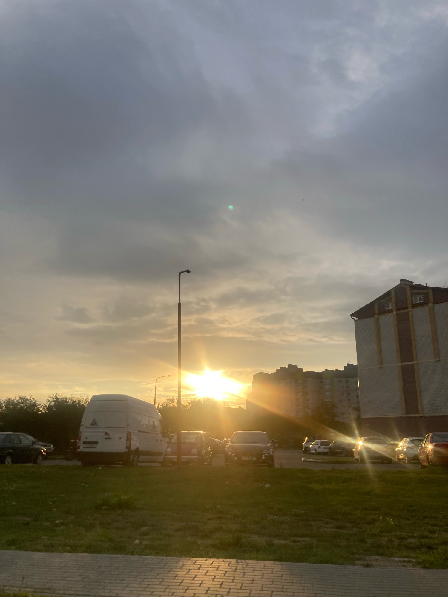 The image shows a street scene at dusk in the city of Brest, Belarus. The sky is overcast with scattered clouds, and the sun is setting, casting a warm, golden light across the scene. Several buildings are visible, including apartment blocks and a taller, brick-clad building. A white van is parked in a grassy area, with several cars parked in the background. There are two streetlights visible, one of which is taller than the other. The ground in the foreground appears to be a paved walkway. The overall impression is a quiet residential area at the end of the day.