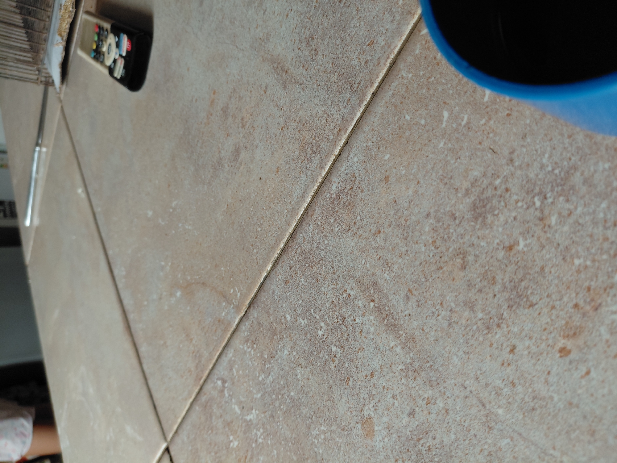 The image presents a close-up, slightly tilted overhead view of a tiled surface, likely a floor or countertop, in an indoor setting in Santa Ana, Venezuela. The surface is covered with light beige or tan ceramic tiles featuring a textured, mottled pattern with subtle variations in shades of brown, grey, and off-white. Thin lines of grout separate the tiles.
In the upper left portion of the frame, a black remote control is visible. It has numerous buttons of various colors, including red, green, yellow, blue, black, and white, with white text or symbols. Icons such as a house, a star, and a circular arrow are discernable, suggesting it's a remote for a modern television or streaming device. Adjacent to the remote, towards the far left edge, blurry details of what appears to be a stack of items and a thin, reflective metallic object are partially visible.
Towards the upper right, a vibrant blue cup or mug is partially captured, with its dark interior suggesting it contains a dark liquid, possibly coffee or tea. The lighting is soft and diffuse, indicating an indoor environment, likely during the daytime. No people are present, and no particular activity or event is taking place; the scene depicts static, everyday household items. No explicit emotions are conveyed, and no discernible text apart from the symbols on the remote control is clearly visible. The overall impression is one of a casual, domestic moment.