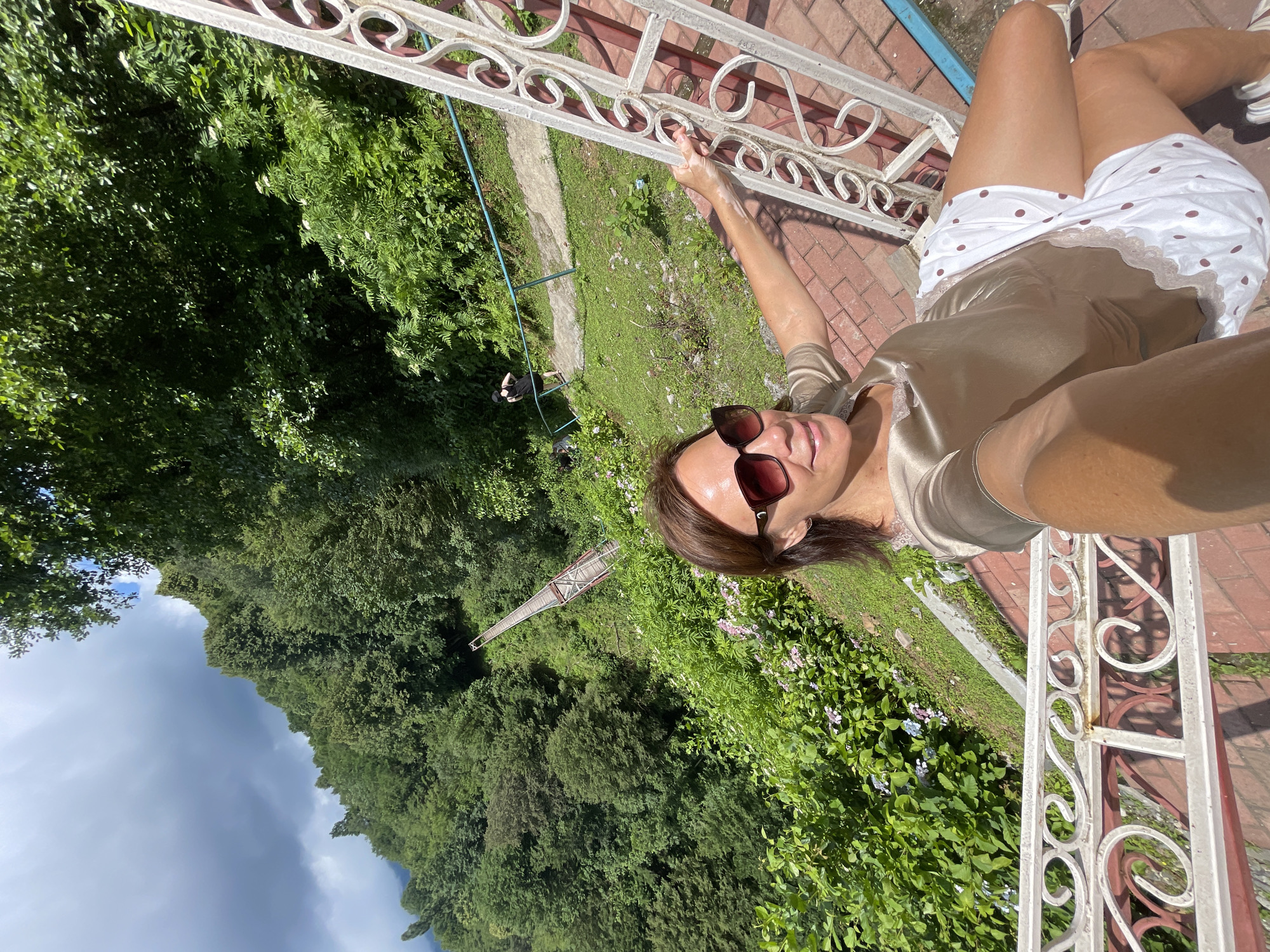 In Tqwarchal, Abkhazia, a woman is captured in a selfie on a red brick walkway with ornate white railings. She is wearing sunglasses, a bronze-colored silk top, and white shorts with brown polka dots. Behind her, lush green trees cover a steep mountainside. A narrow wooden bridge with red supports spans the verdant slope in the distance. Further back, two other individuals are visible on a pathway beside the trees, with one ascending a slope. The sky above is a mix of white clouds and blue.