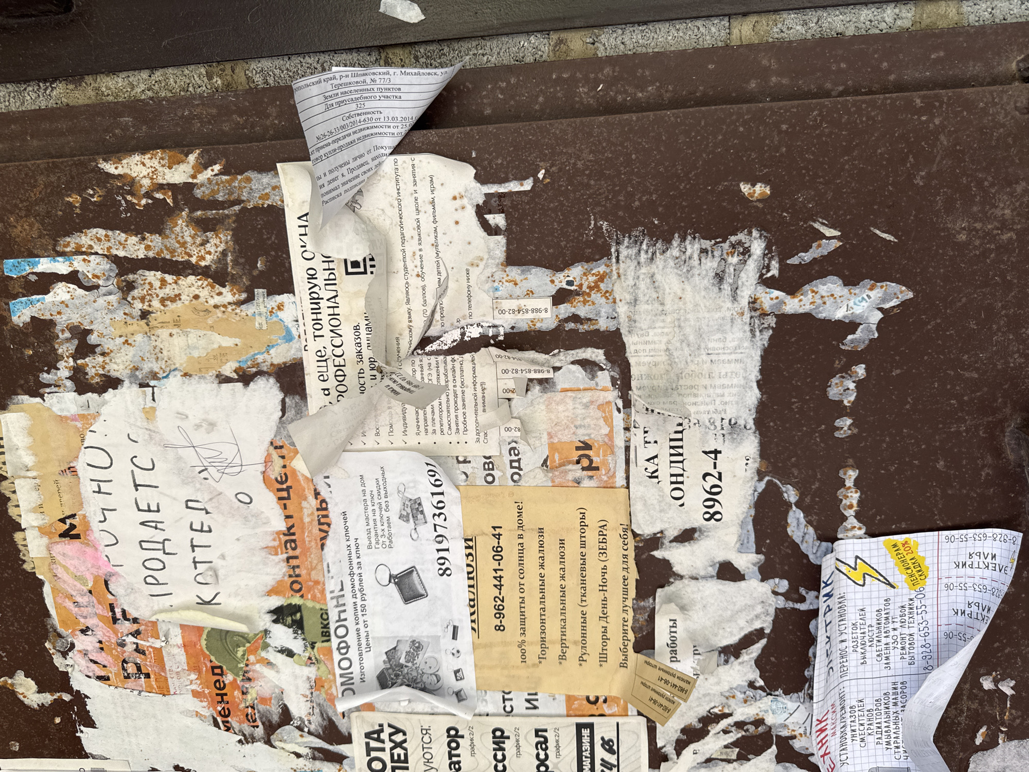 The image shows a close-up, eye-level, outdoor, full shot of a rusty metal surface covered with numerous torn and overlapping advertisements and flyers. The advertisements are printed in Russian and feature various services including locksmiths, window cleaning, and electrical repairs. Some of the flyers are peeling off, revealing the weathered texture of the metal underneath. The overall scene suggests a public bulletin board or a wall used for posting advertisements in a city. One advertisement in the lower right corner mentions "г. Михайловск", indicating the location of Mikhaylovsk, Russia. The image is well-lit, and the details of the flyers, including text and some small illustrations, are visible despite the overall wear and tear.