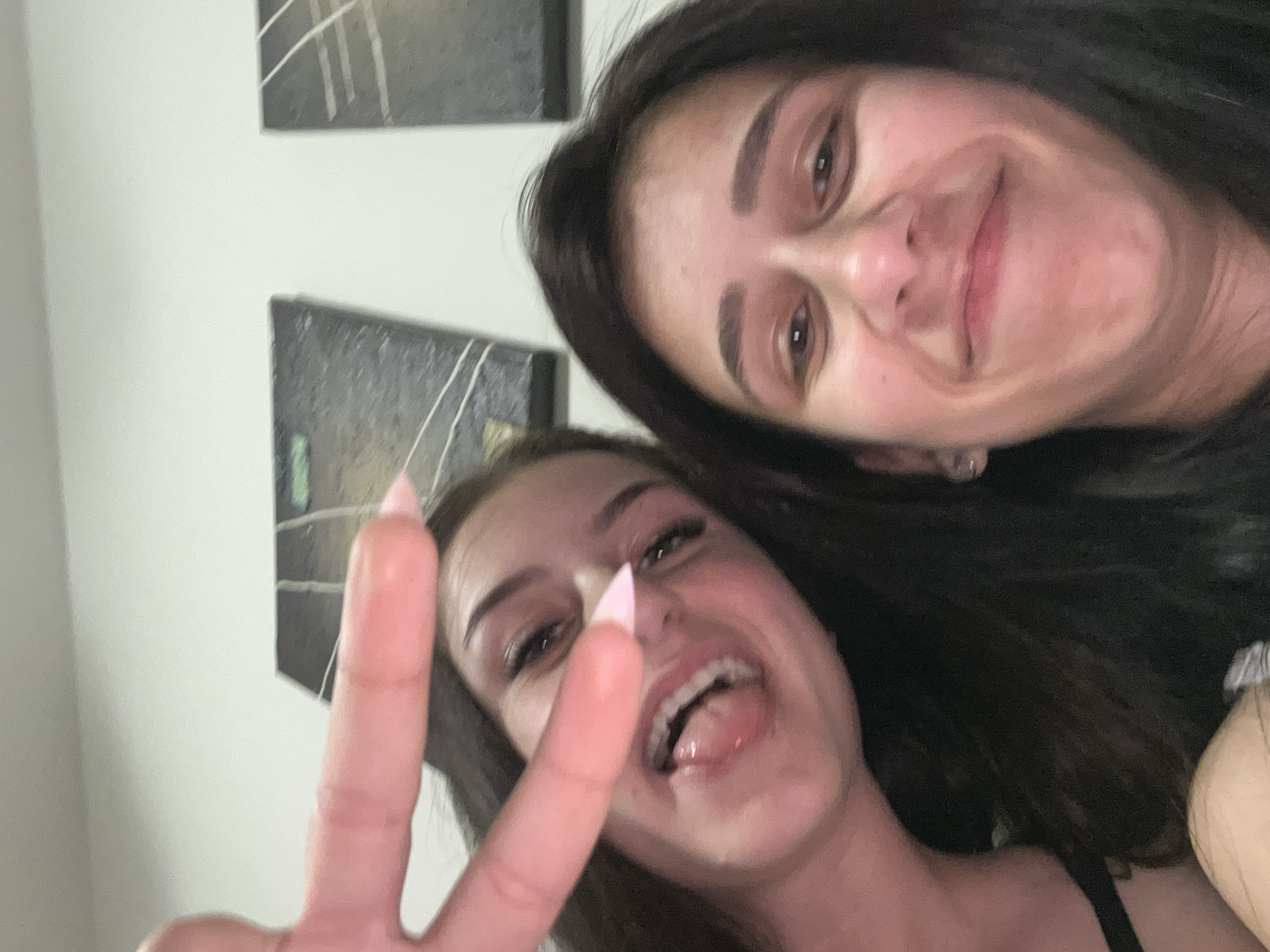 Two women are posing for a selfie indoors. The woman in the foreground is flashing a peace sign and has her mouth open. Both women have dark hair. Paintings with white lines decorate the wall behind them. There is no information that would indicate this is in Plovdiv, Bulgaria.