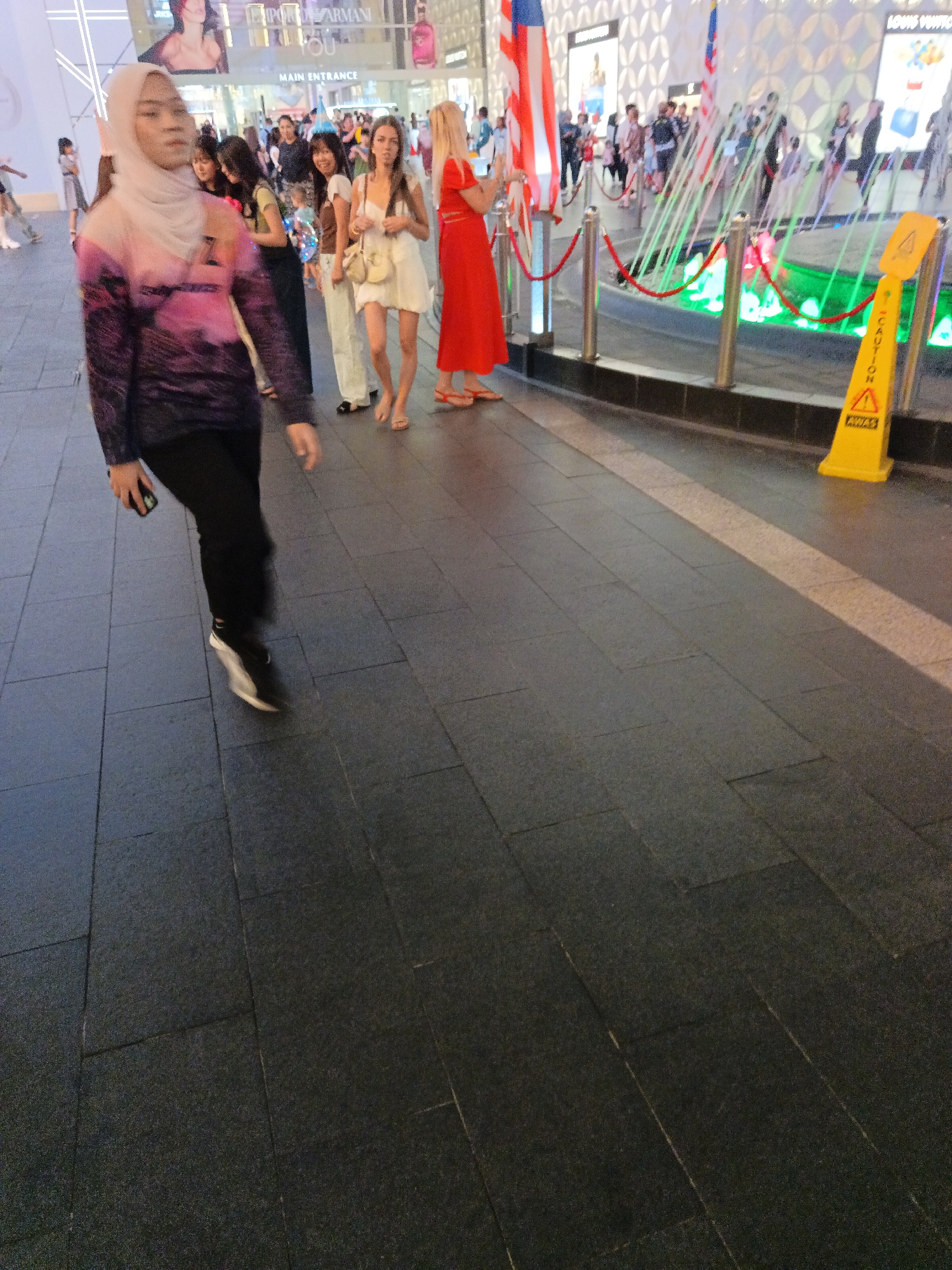 The image depicts a street scene at night in Kuala Lumpur, Malaysia, likely in the vicinity of a commercial area given the presence of illuminated storefronts and signage. In the foreground, a woman wearing a hijab and a long-sleeved shirt with a landscape design walks towards the left of the frame, holding a mobile phone in her right hand. She is wearing black pants and black and white sneakers. Behind her, a group of people are visible, including several women in casual attire, some appearing to be tourists or shoppers. One woman in a white dress and a straw hat is looking towards the camera, while another in a red dress stands further back. A yellow caution sign with a wet floor symbol is positioned on the right side of the image, indicating potential hazard on the tiled pavement. The background reveals the façade of a modern building with prominent signage, including what appears to be "MAIN ENTRANCE." Decorative lights and water features are also visible, suggesting a lively urban environment. The overall lighting suggests it is evening or nighttime, with artificial lights illuminating the scene.