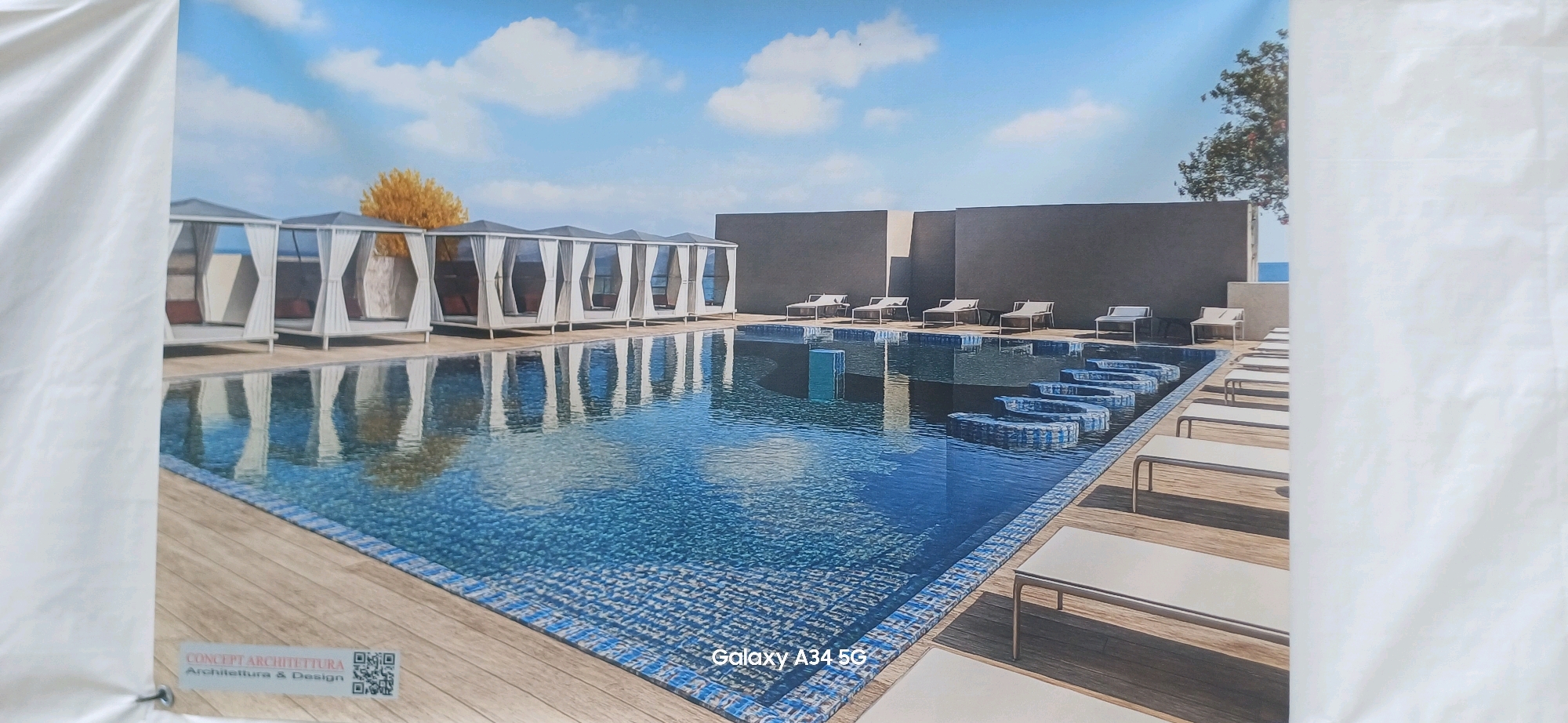 This is a digitally rendered image of a modern outdoor swimming pool area. The pool itself is large and irregularly shaped, with a curved section featuring built-in seating or steps. The water is a deep blue, reflecting the sky and surrounding structures. The pool deck is made of light-colored wooden planks.
To the left of the pool, there are several white, open-sided cabanas with draped white curtains, each containing what appears to be a daybed or lounge seating. Behind these cabanas, a solitary tree with golden foliage stands out against a bright blue sky with scattered white clouds.
In the background, to the right of the cabanas, a dark gray wall forms a backdrop to a row of white lounge chairs situated on the pool deck. Further to the right, a portion of a building with light-colored accents is visible, and beyond that, a glimpse of the ocean.
At the bottom left of the image, there is a logo for "CONCEPT ARCHITETTURA Architettura & Design" along with a QR code. In the bottom right corner, the text "Galaxy A34 5G" is displayed.
The image does not contain any people or animals. The scene depicts a luxurious and serene environment, likely a resort or a high-end residential property.
Regarding the location in Abano Terme, Italy, this image does not contain any specific visual cues or text that would definitively place it in Abano Terme. The provided information indicates this is a rendering, and without further context or metadata, its specific real-world location cannot be determined from the image alone.