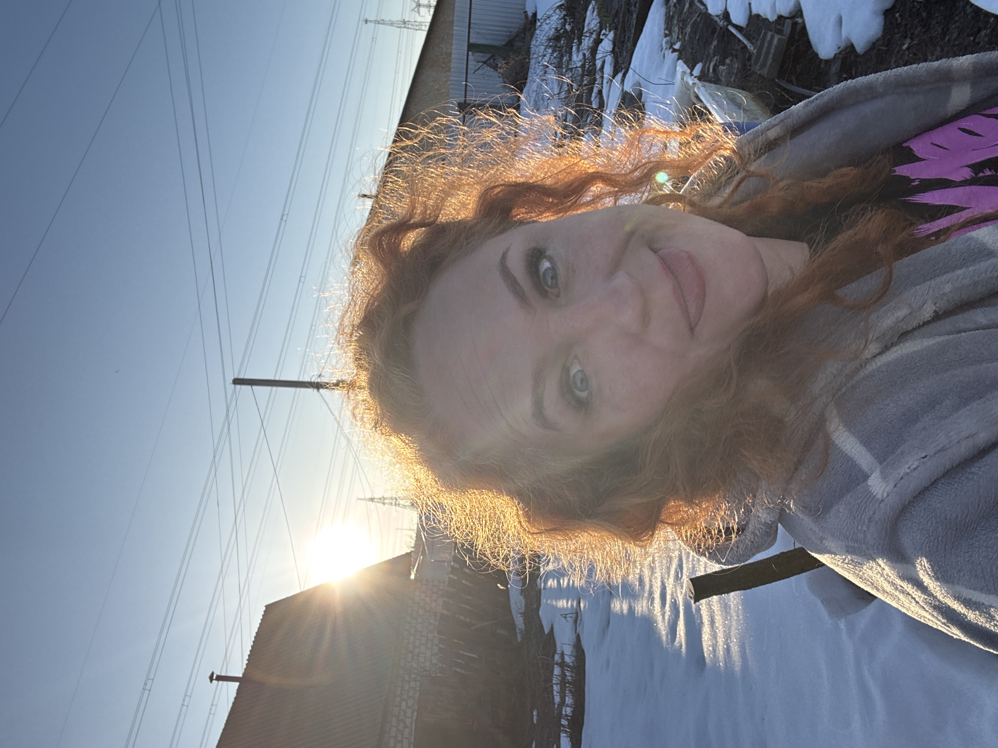 A woman with fair skin and red hair is taking a selfie outdoors. She is wearing a grey jacket with white trim and a purple and black patterned shirt underneath. The sun is behind her, creating a halo effect around her hair. In the background, there is a building with a sloping roof, power lines against a blue sky, and patches of snow on the ground. The location is likely Chaikovsky, Russia based on prompt context.