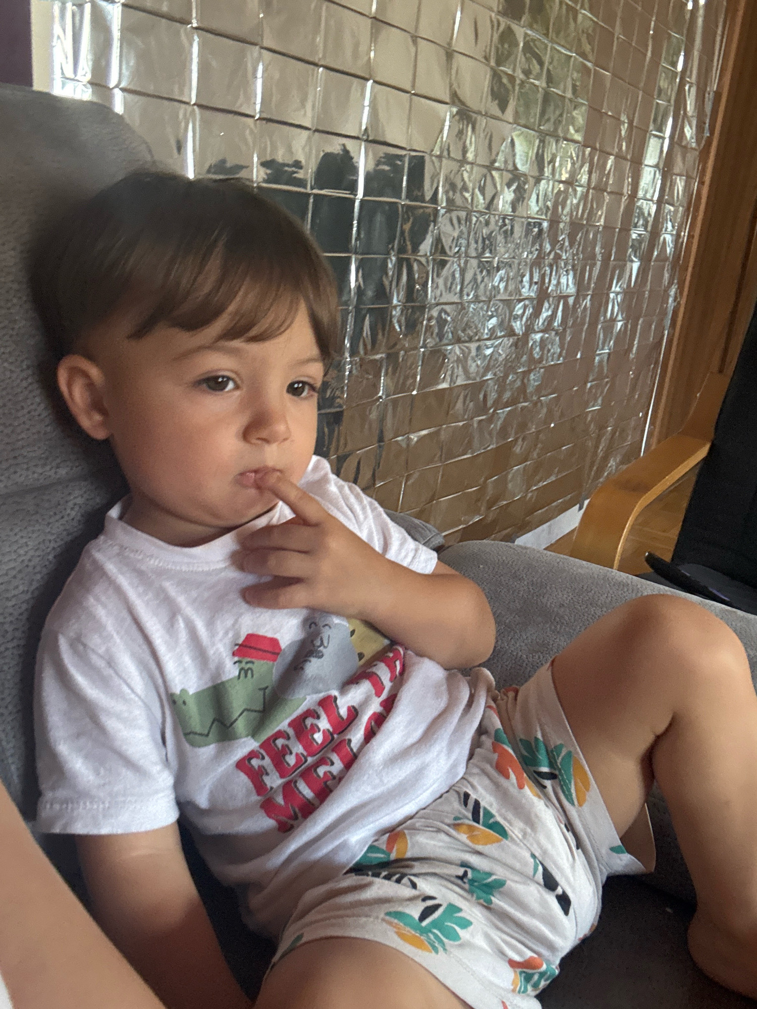 A young boy with brown hair and brown eyes is sitting in a gray armchair, resting his chin on his hand. He is wearing a white t-shirt with a graphic of a crocodile and the words "FEEL ME" printed on it. He is also wearing light-colored shorts with a leaf pattern. The background is a wall covered in reflective material, possibly insulation, and a wooden chair is visible to the right. The image appears to be taken indoors, possibly in a home in Überherrn, Germany.