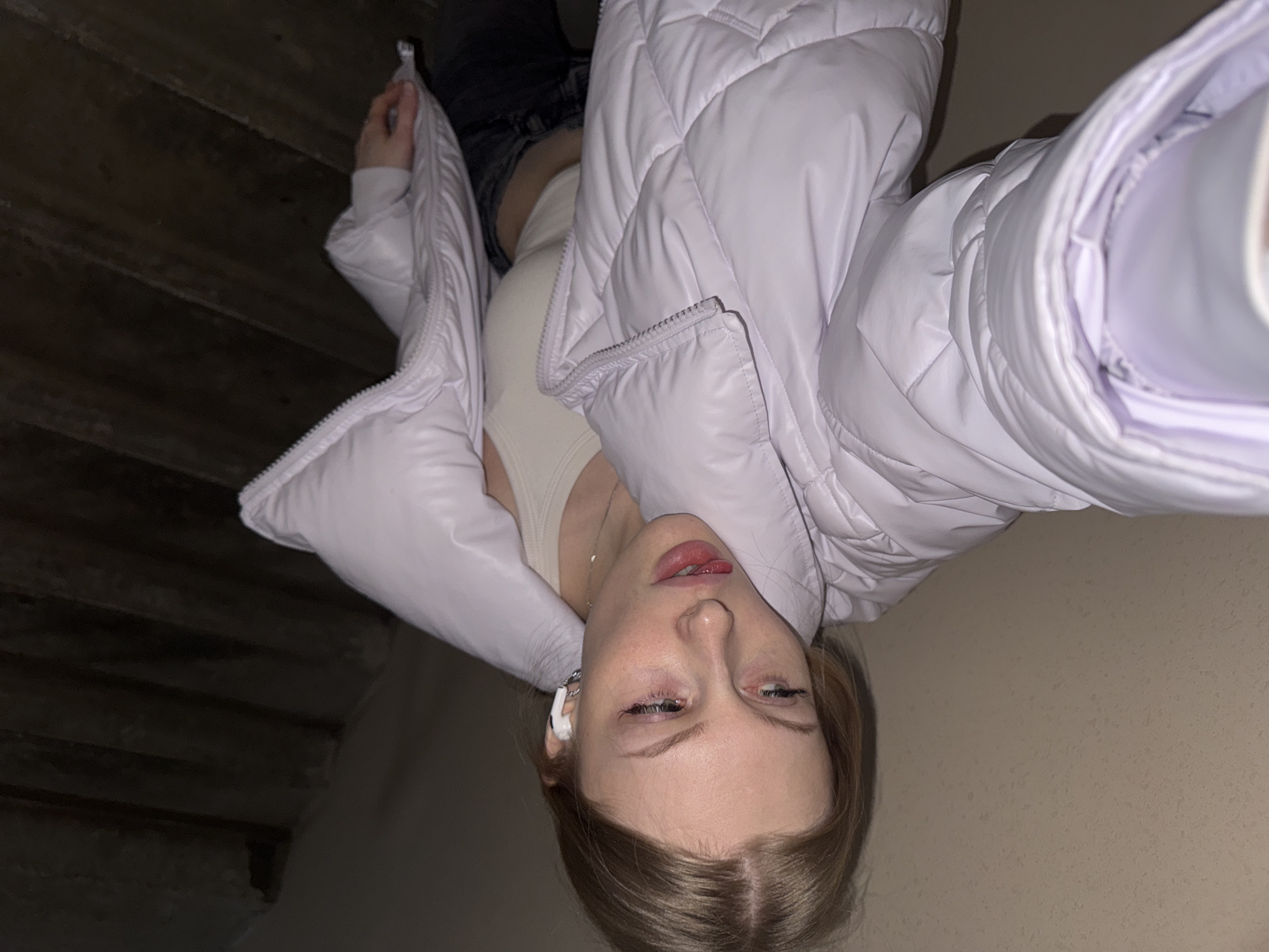 A young woman is captured in a selfie, taken indoors in what is presumably Perm, Russia. She is positioned centrally in the frame, viewed from the upper torso up, looking directly at the camera with her head tilted. She is wearing a light lavender or very pale purple puffer jacket, unzipped to reveal a cream-colored top beneath. A slender silver necklace is visible on her neck, and a white earbud is inserted in her left ear.
Her expression is casual, with slightly heavy-lidded eyes and a partially open mouth where her tongue subtly protrudes, suggesting a relaxed or possibly playful mood. Her left arm, which appears to be holding the camera, is visible on the right side of the frame.
The background on the left features a dark, textured surface with horizontal lines, strongly resembling wooden stairs or steps in a dimly lit environment. On the right, there is a plain, light beige wall. The lighting throughout the image is dim, indicating an indoor setting with low ambient light, potentially suggesting evening or nighttime. No other subjects or distinct objects are present, and there is no visible text in the image.