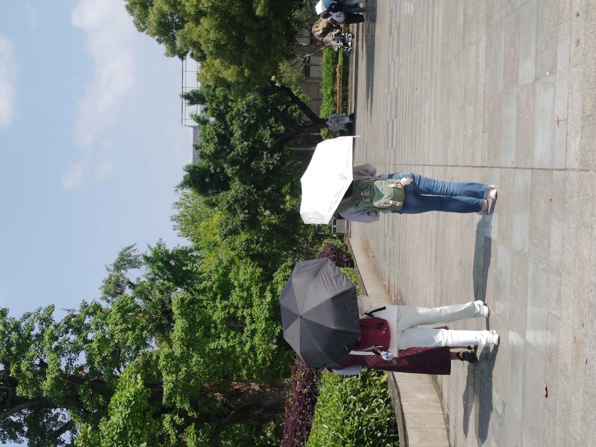 The image, taken in Chongqing High-tech Zone, China, depicts an outdoor scene on a bright, sunny day. Two individuals are prominently featured in the foreground and midground, walking away from the viewer on a wide, light-colored paved walkway.
The person in the foreground is holding a black umbrella, providing shade. They are wearing a light-colored top, a dark red backpack, and a plaid skirt (appearing red and light-colored, possibly white or light blue). Their legs are visible below the skirt, wearing light shoes, and a distinct shadow stretches out behind them on the pavement.
Slightly further away, another person is also walking away from the camera, holding a white umbrella for sun protection. This individual is dressed in a light-colored top, blue jeans, and carries a green backpack. Their shoes also appear light-colored, and a shadow is cast on the ground.
The paved walkway, made of rectangular tiles, extends into the distance, with a few scattered leaves on its surface. To the left of the path, lush green trees with dense foliage create a natural border, accompanied by some darker, reddish-purple bushes. In the background, beyond the trees, hints of urban structures or buildings are visible under a clear, bright blue sky dotted with a few scattered white clouds. The strong sunlight and the use of umbrellas suggest it is midday or early afternoon, and likely warm or hot weather. Several other indistinct figures can be seen in the far background, some appearing to be sitting or standing near trees. No visible text is present in the image.