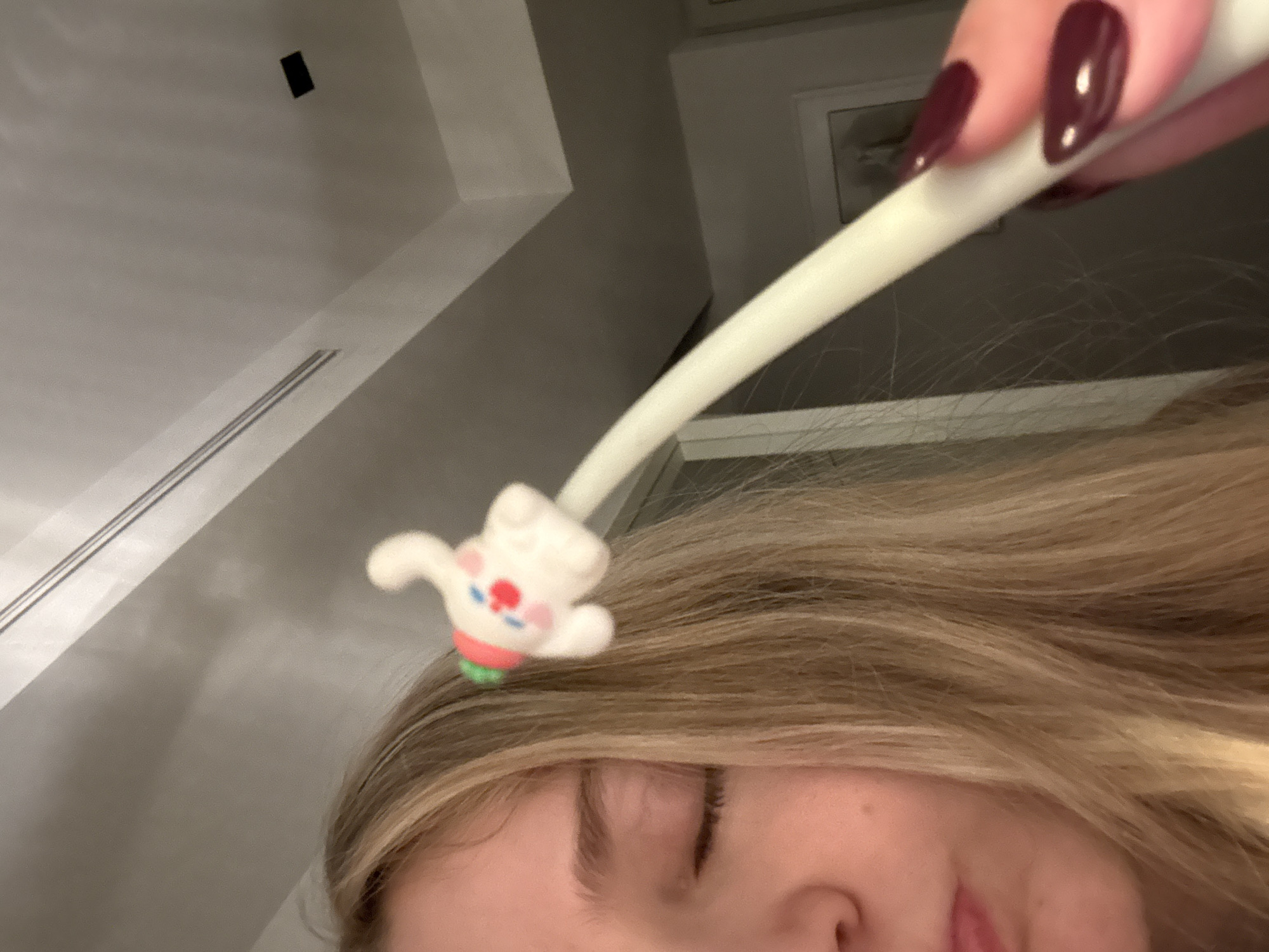 A close-up, low-angle shot focuses on a person, likely a woman, with blonde hair and closed eyes. A hand with dark-colored, manicured nails holds a white, curved object with a small, fuzzy, white animal attached to the end over the person's head. The animal has a red and green design. The background consists of a white wall with a linear vent and a darker wall with a framed object. The location is likely indoors. No specific details indicate it is in Mexico City, Mexico.