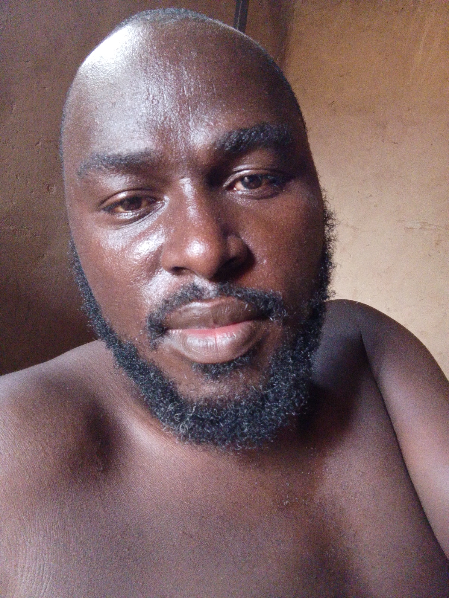 A medium close-up photograph depicts a dark-skinned, bald Black man with a short, dark beard. His face is shiny, possibly due to sweat or oil. He gazes directly at the camera with a somber expression. The background appears to be a nondescript wall of a tan or light brown color, possibly painted or textured. The man's bare chest and shoulders are visible in the foreground. The location is unknown.
