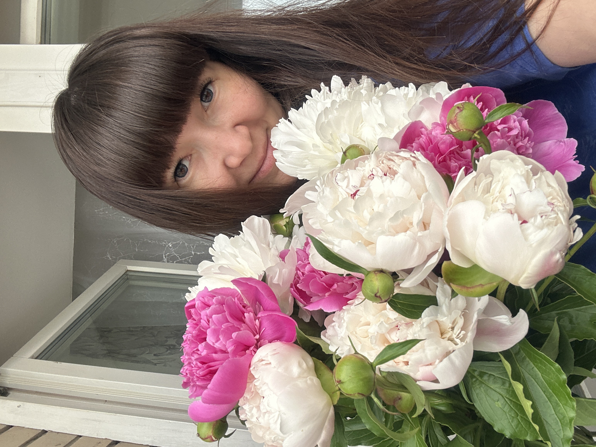 A woman with brown hair and bangs is holding a bouquet of peonies. The bouquet contains white and pink peonies with green leaves and buds. She is smiling slightly at the camera. Behind her is a window with a white frame. The location is implied to be in the city of Chaikovsky, Russia.