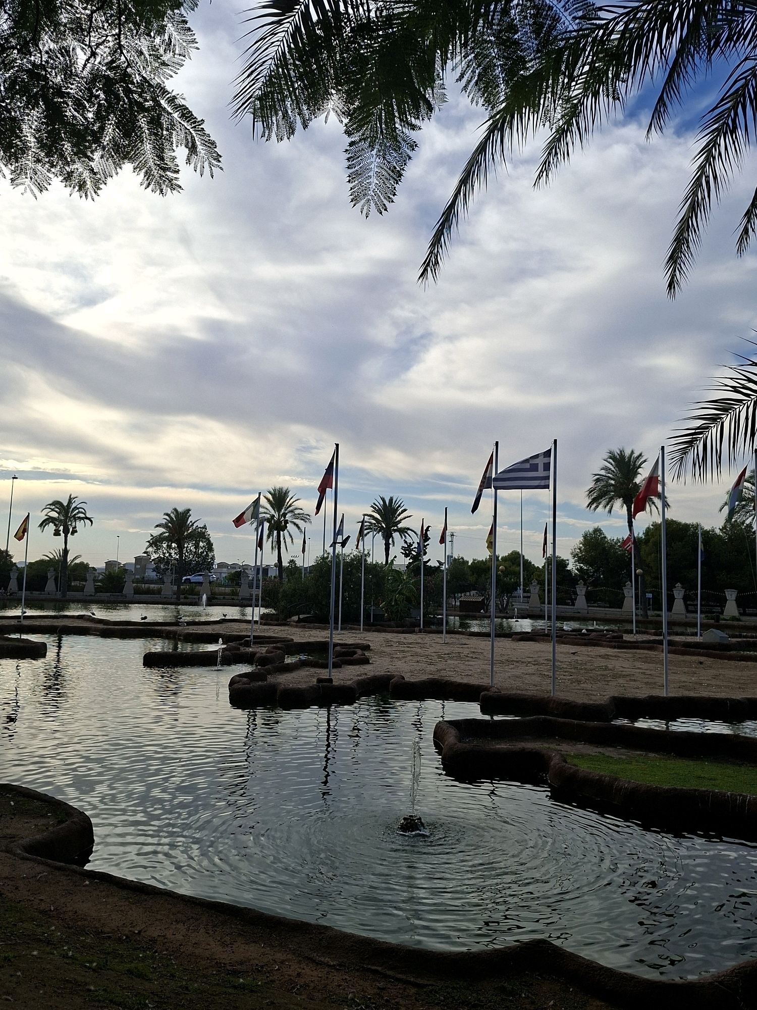 The image depicts a park-like setting with a pond in the foreground, featuring several small fountains. The pond's edges are lined with dark, irregular borders and some areas of green grass. Beyond the pond, a sandy area is populated with numerous flags from different countries displayed on tall poles. In the background, palm trees and other vegetation are visible, with buildings and infrastructure further in the distance. The sky is partly cloudy, with a soft, diffused light suggesting either early morning or late afternoon. The scene is tranquil, with no people or specific actions clearly identifiable. This appears to be a representation of the "Parque de las Naciones" (Park of the Nations) in Torrevieja, Spain, which is known for its display of flags from various countries.