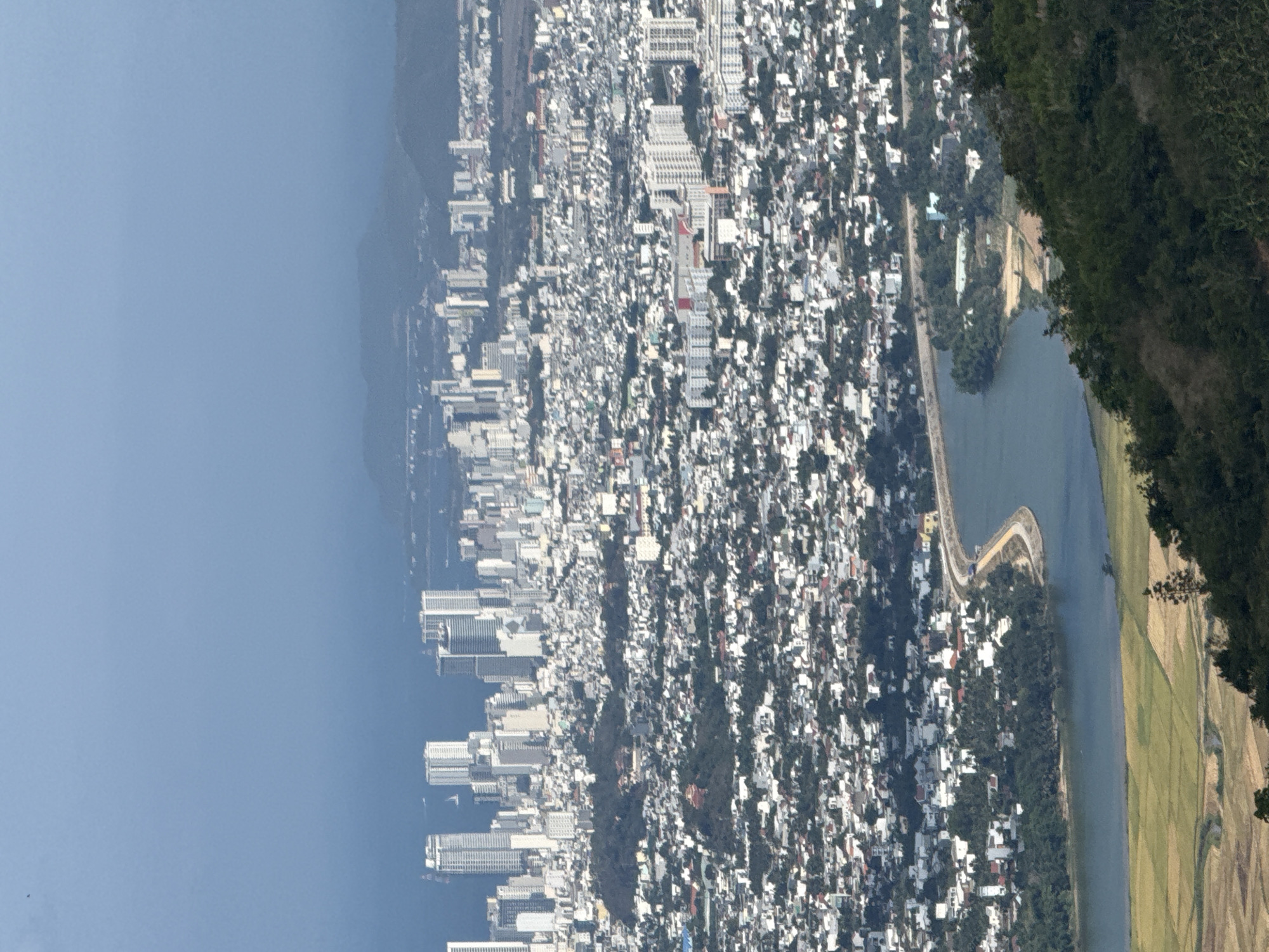 The image displays a high-angle, expansive view of a coastal city and its surrounding landscape under clear, daytime conditions.
On the left, a large expanse of serene blue water, likely a sea or bay, stretches towards the horizon. Adjacent to this water body, a dense urban environment unfolds, characterized by a multitude of buildings. Closer to the waterfront, several modern, multi-story buildings and high-rises are visible, suggesting a commercial or densely residential district. Further inland, the city appears to consist of a mix of smaller, clustered structures and some interspersed green areas, indicative of a sprawling urban development.
A river or channel winds through the scene, separating parts of the urban area from more natural landscapes. A thin strip of land or a road/dam structure can be seen curving along the bank of this waterway. To the right of the river, agricultural fields, possibly rice paddies, display patterns of green and brown, indicating cultivation. The rightmost portion of the image is dominated by a steep, heavily forested hillside or mountain, covered in dense, dark green foliage, providing a natural boundary to the urban expansion.
The sky is a uniform, clear light blue, with a slight atmospheric haze over the distant water. No specific human activities or individuals are discernible due to the high vantage point and distance. There is no visible text in the image. The scene captures a dynamic landscape where urban development meets natural elements, typical of a coastal city in a tropical region like Tay Nha Trang Ward, Vietnam.