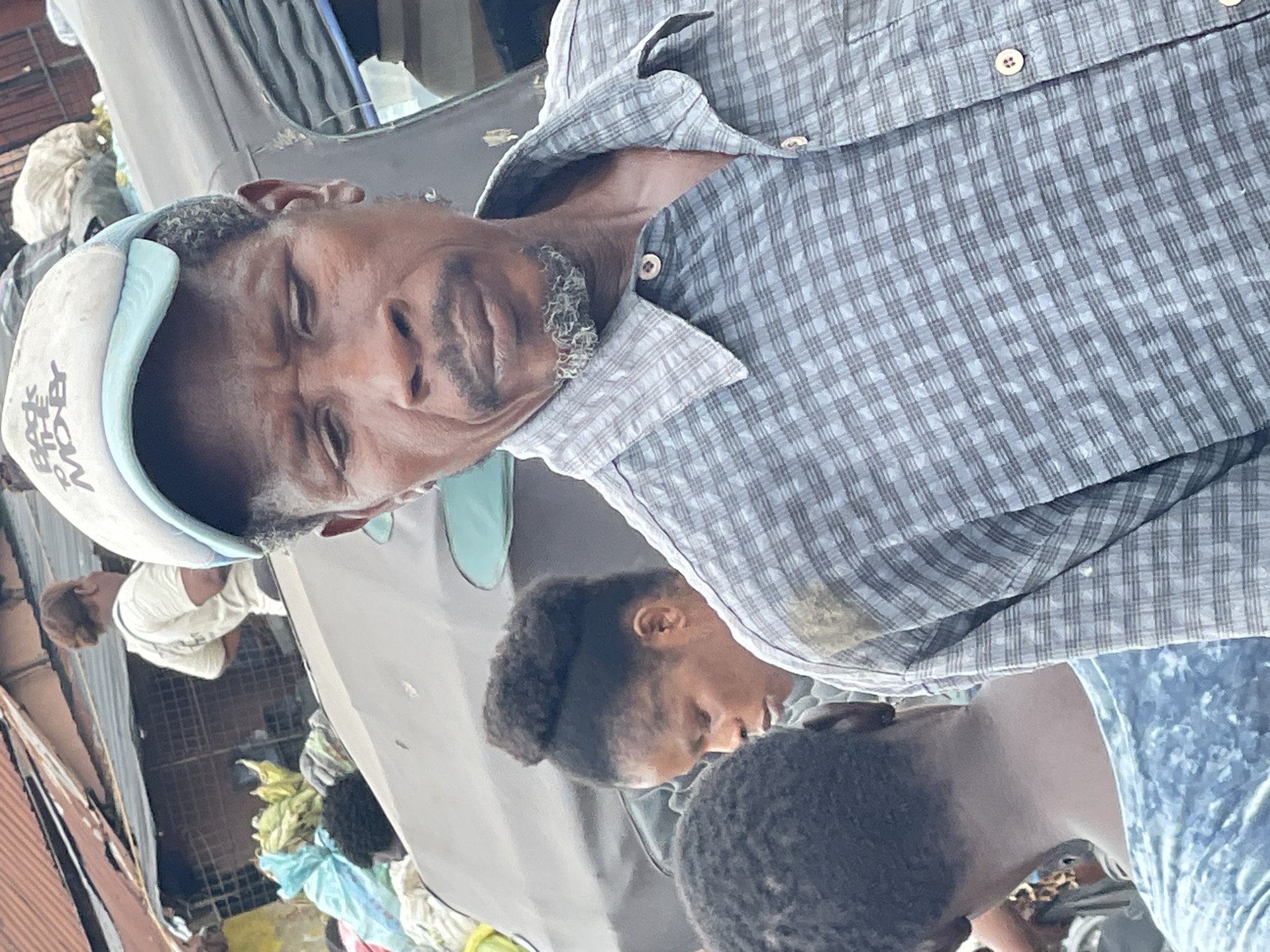 A man wearing a blue and white baseball cap with "BACK TO THE MO BY" printed on it is in the foreground, looking down with his mouth slightly open. He has a goatee and is wearing a plaid collared shirt. To his left and slightly behind him, a woman with her hair in a bun is looking downwards. Behind her, another person's head is visible, facing away from the camera. In the background, a stall with a corrugated metal roof and a variety of items, including what appears to be fabric and bags, is present. The scene appears to be an outdoor market in Fegge, Nigeria, with a vehicle partially visible in the background.