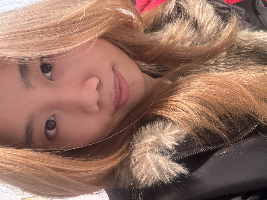 An adult East Asian female is depicted in a close-up, head-and-shoulders portrait, observed in Shapingba District, China. The individual has light brown, wavy hair that cascades around her face and shoulders. Her dark brown eyes are open, focused directly on the camera. Her lips are closed, presenting a neutral or subtly smiling expression. She is wearing a dark-colored jacket, likely black, featuring a prominent faux fur collar or hood lining in shades of brown and beige. A silver-toned metal zipper is visible on the jacket's front. A small portion of a red garment is discernible on the far right of the frame, partially obscured by her hair and jacket. The background is out of focus and appears brightly lit, devoid of discernible environmental features.