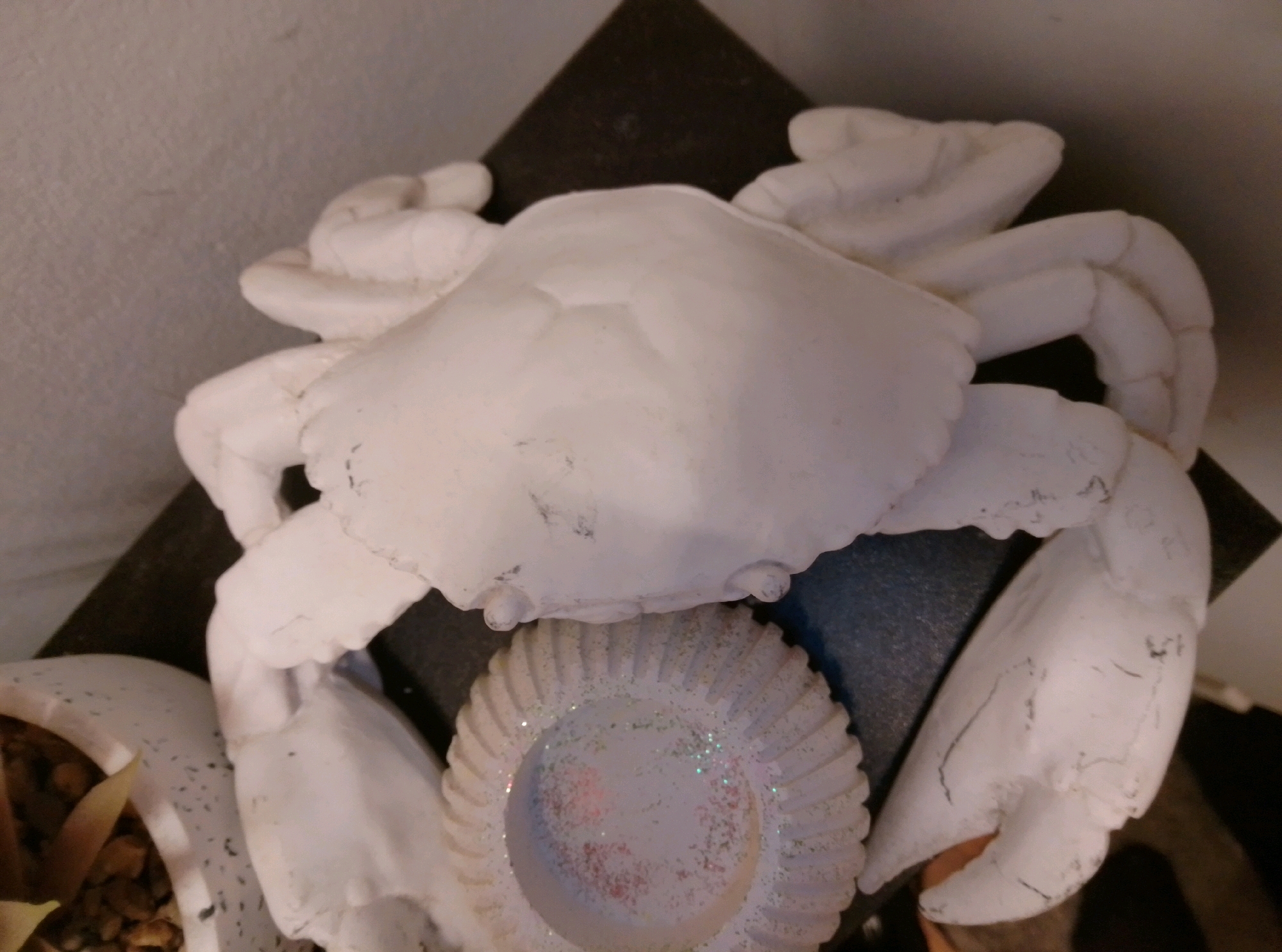 A white plaster crab sculpture rests on a dark triangular surface, which is itself placed on a dark textured surface. To the left of the crab sculpture, a portion of a white planter with gravel and a fallen leaf is visible. The crab sculpture is centrally located and appears to be the primary subject. A decorative ridged circular object, possibly a candle holder with glitter, is positioned beneath the crab's body. The background consists of a textured off-white wall. The scene appears to be an indoor still life or decorative arrangement. There is no indication of the city of Wedel, Germany, in the image.