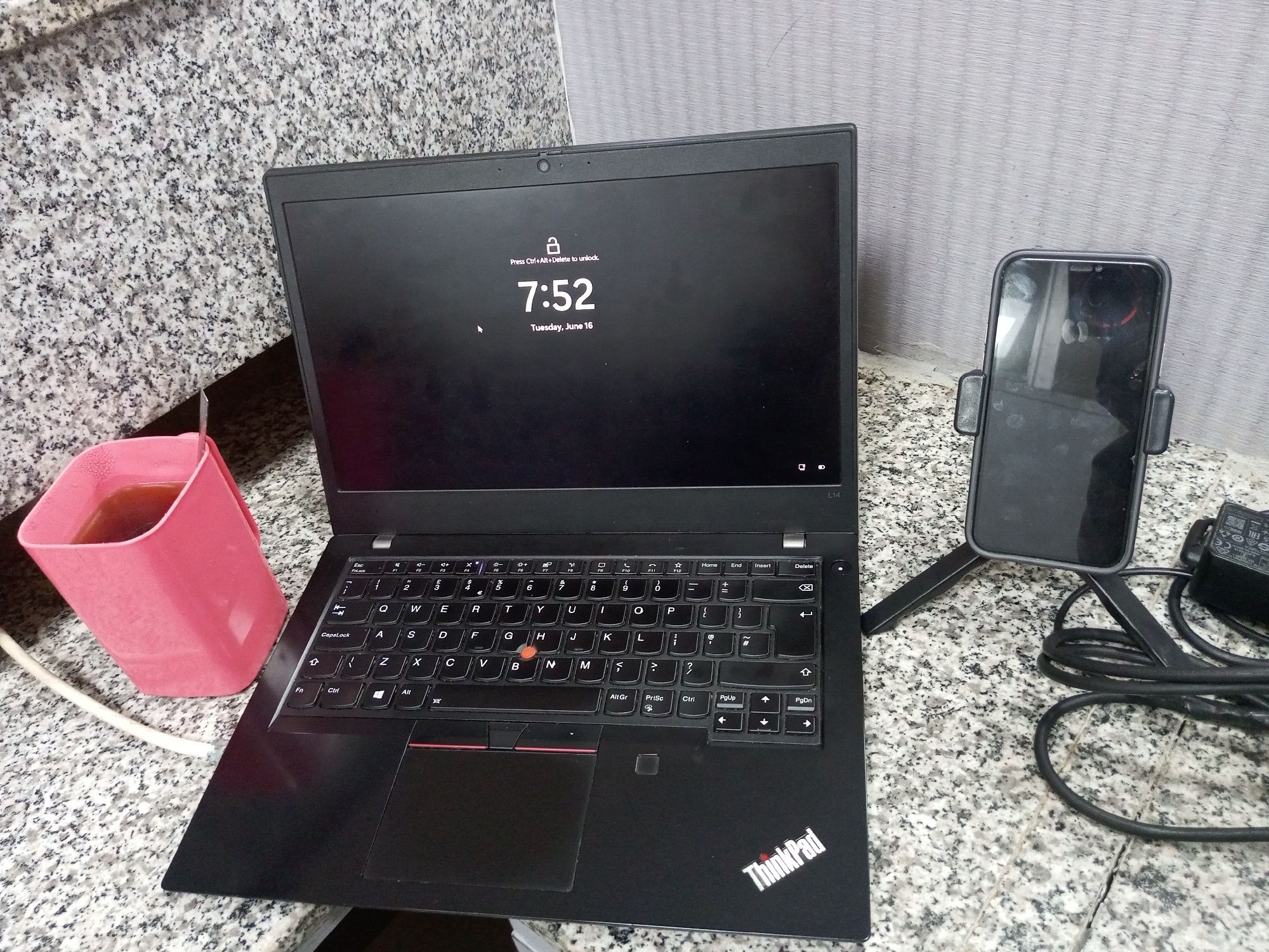 A laptop is positioned on a granite countertop, displaying a locked screen with the time "7:52" and the date "Tuesday, June 16." To the right of the laptop, a smartphone is mounted on a stand. A pink cup filled with a dark liquid sits to the left of the laptop, with a white cord emerging from it. The background shows a tiled wall.