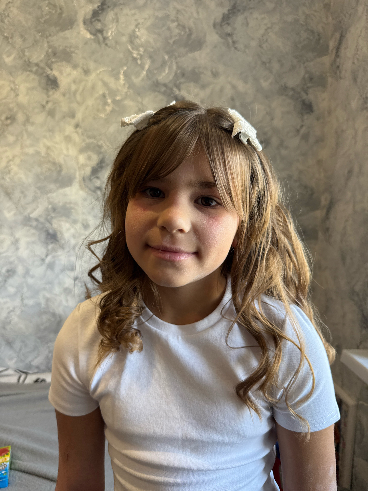 A young girl with long, wavy, blonde-brown hair and bangs, adorned with two white hair clips, is pictured. She is wearing a white t-shirt and has a subtle blush on her cheeks. The background appears to be a textured wall with a marbled or cloudy pattern in shades of grey and white. The lighting suggests an indoor setting, likely a room in a residential dwelling. The girl is looking directly at the camera with a gentle smile. There are no explicit indicators of location within the city of Rusanovo, Russia, present in the image itself.