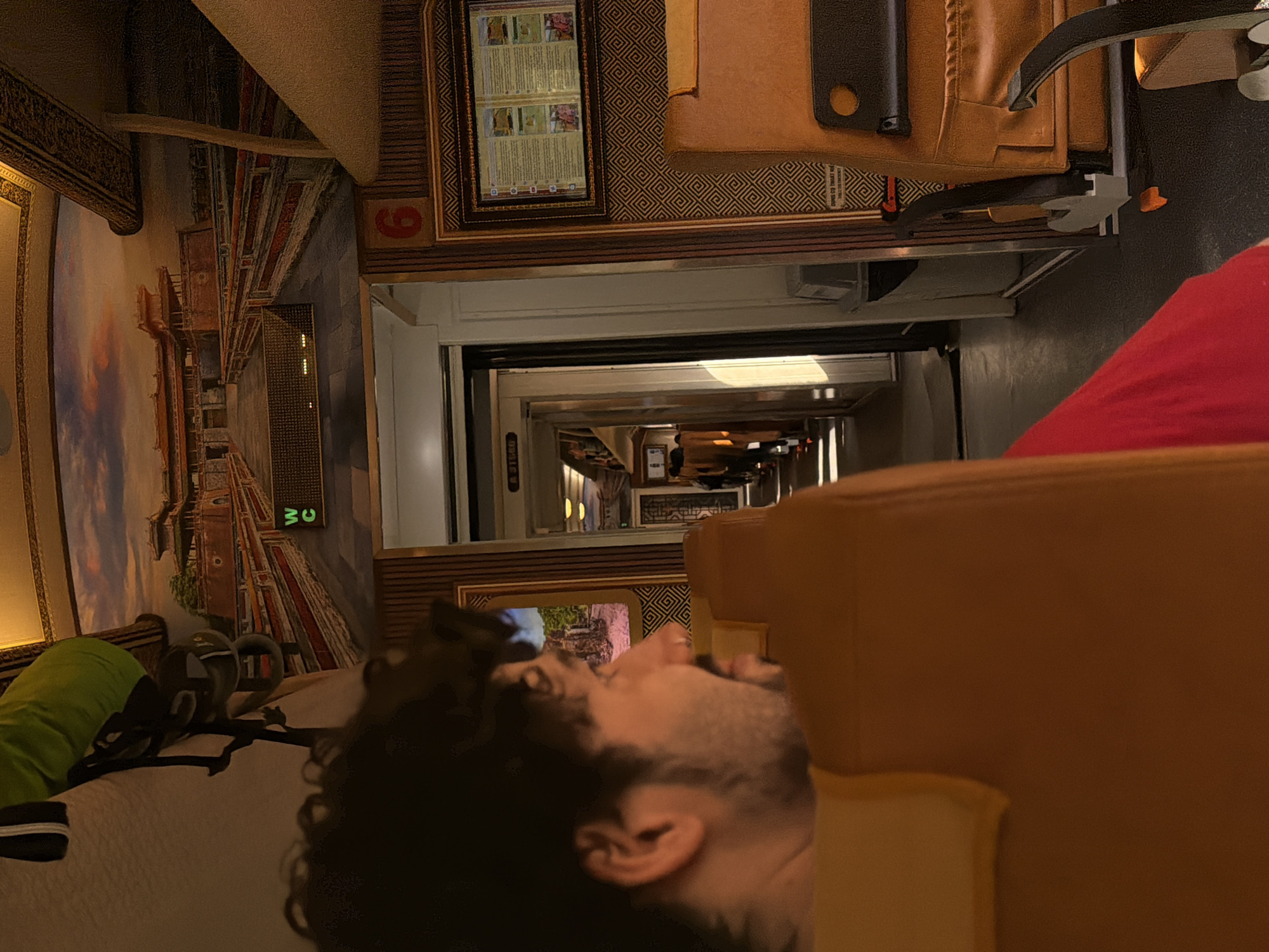 The image shows the interior of a train traveling in Huế, Vietnam. The photo captures a man reclining in a seat, with the train's corridor visible in the background. The train car walls are decorated with images, and a sign indicates the restroom is nearby.