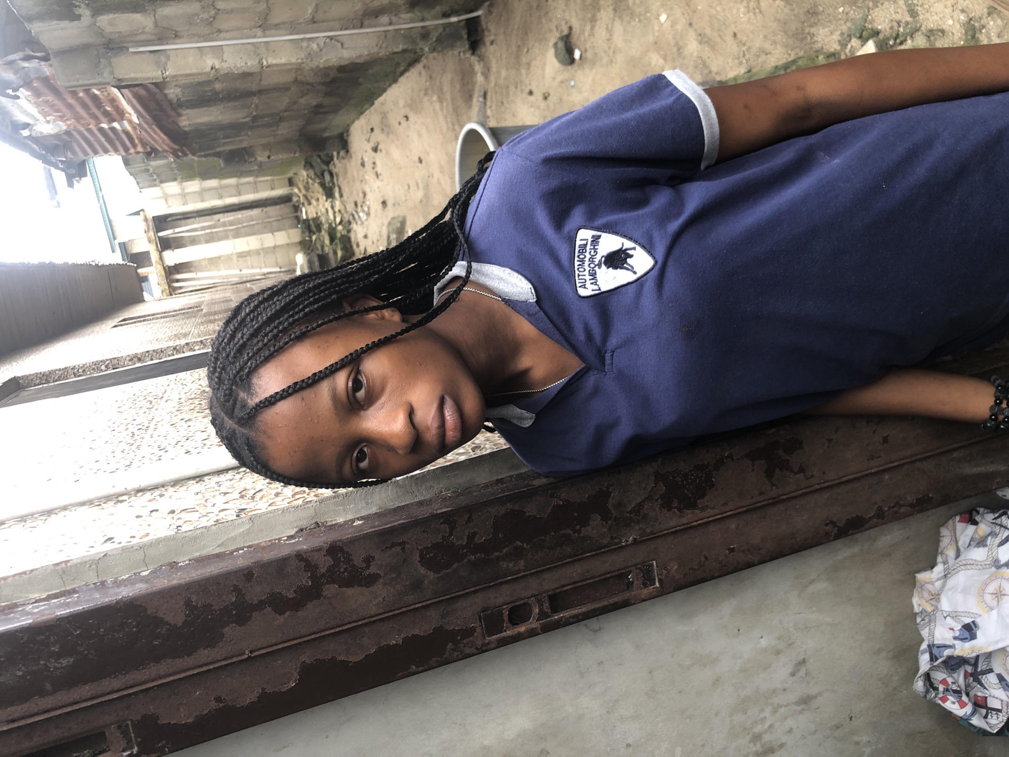 A young Black woman with braided hair lies on her side, her head resting on a weathered, rusty metal surface. She is wearing a dark blue t-shirt with a grey collar and trim. A white emblem with a black silhouette of a bull and the text "AUTOMOBILI LAMBORGHINI" is visible on the shirt. Her expression is neutral.
The setting appears to be an outdoor, possibly urban or semi-urban environment in Ado, Nigeria. In the background, there are concrete structures, corrugated metal sheeting, and rough ground. The lighting suggests it is daytime. A white cloth with a nautical print is partially visible in the lower right corner of the frame. The overall impression is a candid moment captured in a somewhat utilitarian or outdoor space.