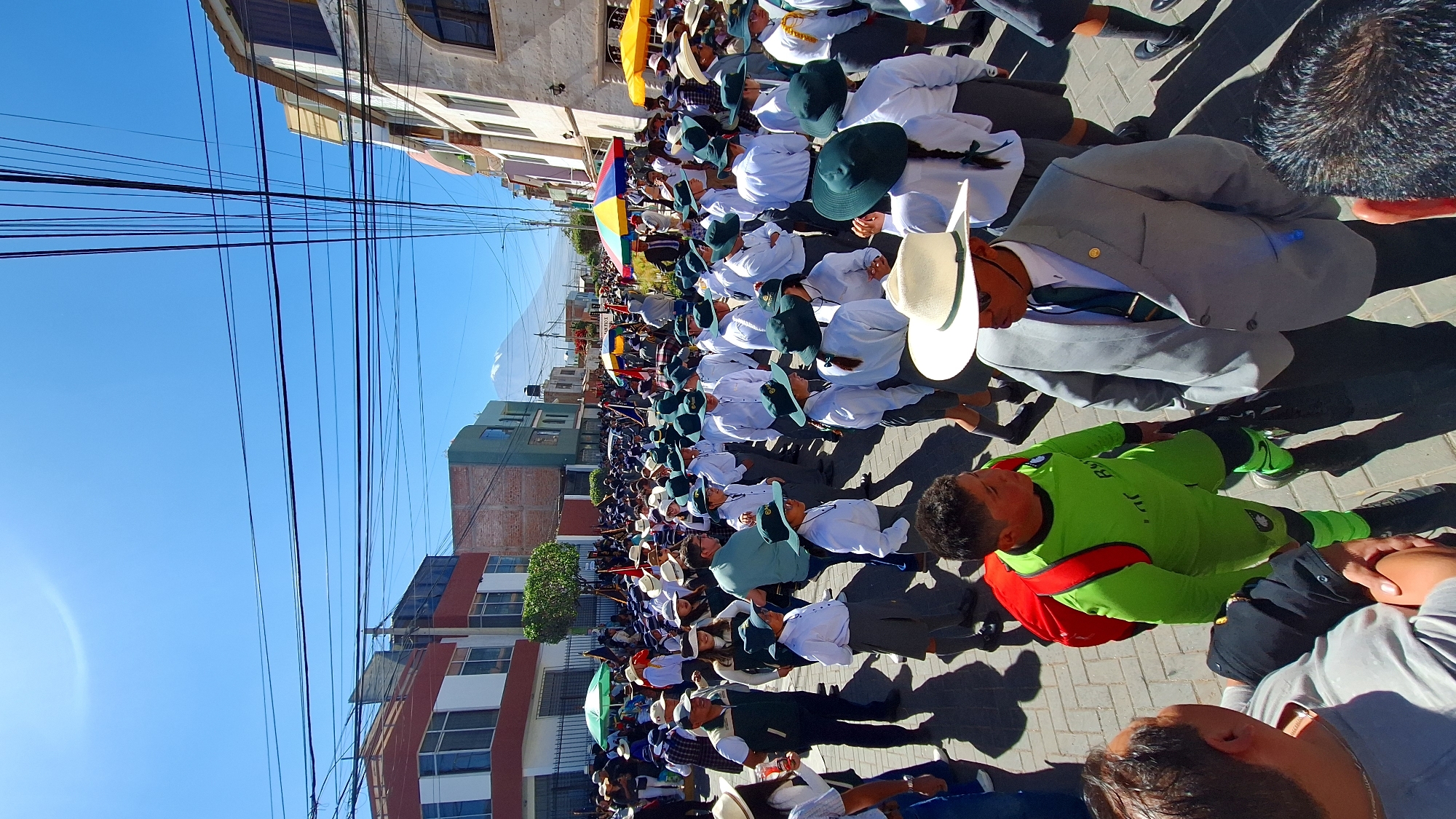 The image depicts a large group of people gathered on a paved street in Cerro Colorado, Peru. The street is lined with buildings, some with multiple stories. Overhead, a network of power lines crisscrosses the sky. Many individuals in the crowd are wearing matching uniforms consisting of white shirts, hats, and dark green accents. One person near the front is wearing a bright green outfit with red accents and another in a grey suit with a white hat. A colorful umbrella is visible in the midst of the crowd. The weather appears to be sunny and bright. The crowd seems to be part of a parade or public gathering.