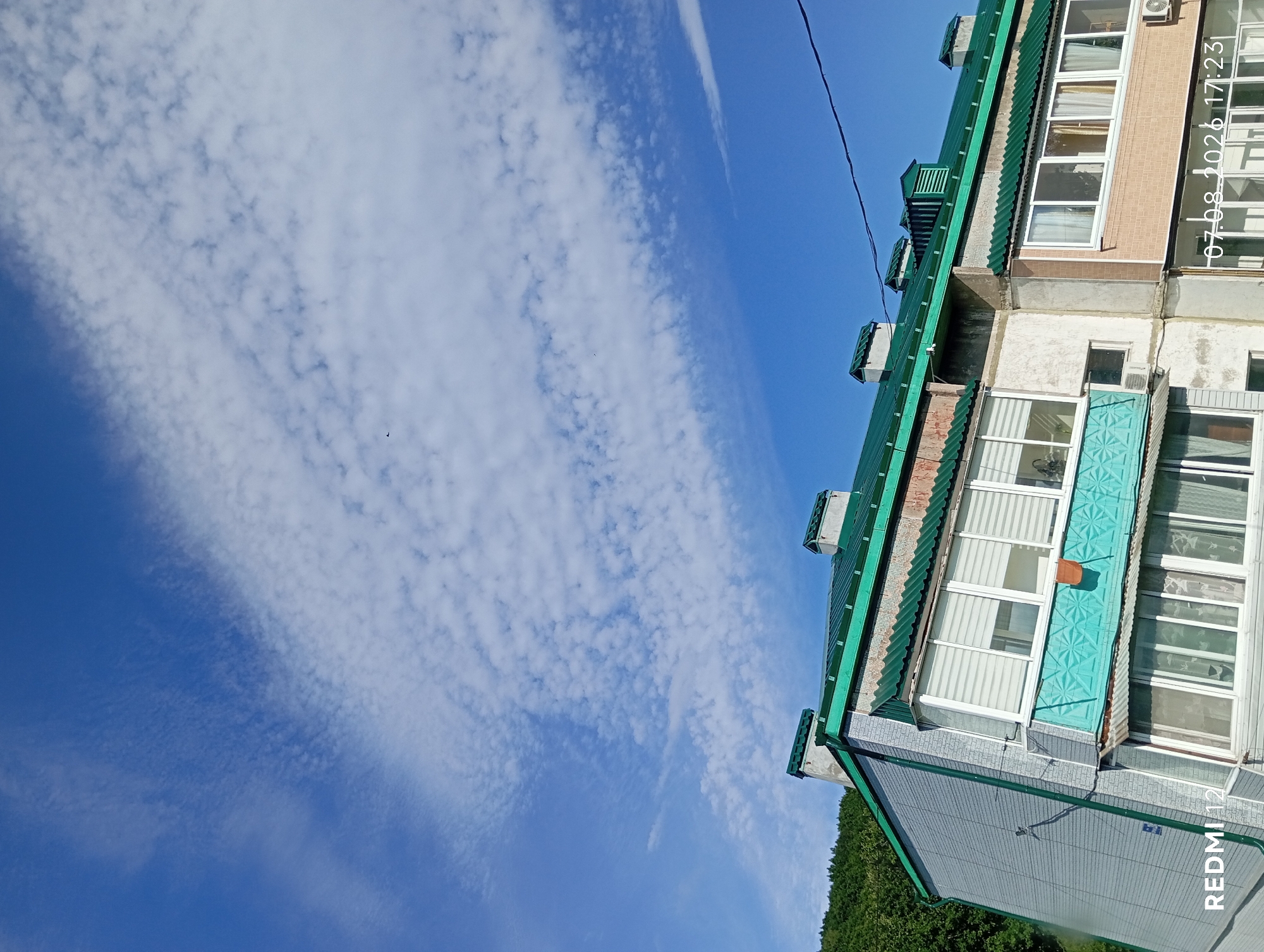 The image shows the upper floors of an apartment building with a green corrugated metal roof and several chimneys. The building has a light gray textured facade on the left, with a light beige brick-patterned facade on the right. There are multiple balconies with white window frames and blinds. A hillside covered with green trees is visible in the background on the left. The sky is blue with scattered white clouds. A date and time stamp "07.08.2026 17:23" and "REDMI 12" are present in the lower corners of the image. The scene appears to be captured from a slightly elevated perspective, looking up at the building. There are no people or vehicles visible in the image. The location is identified as Baranovskiy, Russia.