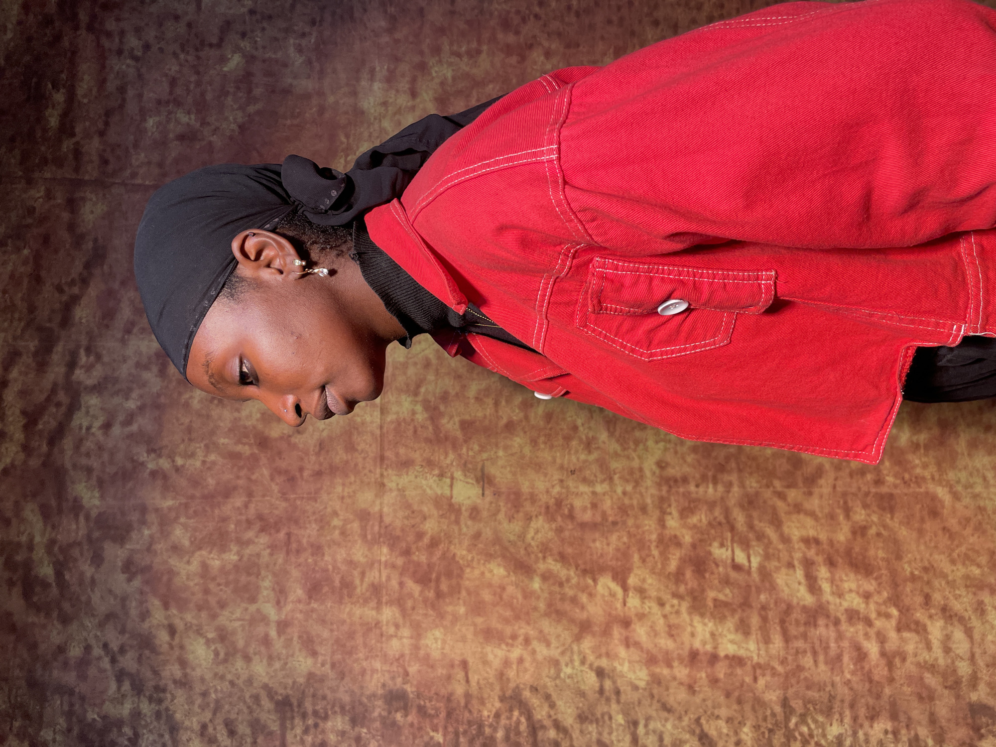 A woman with dark skin, wearing a black head covering and a red jacket, is positioned in front of a mottled brown backdrop. She is looking down and to the left. Her earring is visible. The location is unspecified, and there is no information to indicate the city of Barakin Akawo, Nigeria.