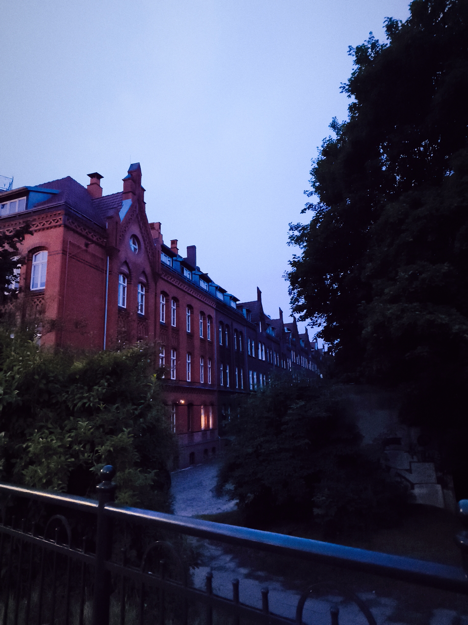 The image depicts the exterior of a red brick building with multiple windows, suggesting it might be an institutional or residential structure. The building features a pitched roof with dormer windows and decorative gables. Adjacent to the building, dense foliage from trees and bushes is visible. In the foreground, a dark metal fence with intricate detailing runs horizontally across the frame. The sky is a pale, twilight blue, indicating either early morning or late evening. The lighting suggests a dim, atmospheric scene. Although the exact location is not specified in the image, the style of architecture and the general environment are consistent with urban settings in Gdańsk, Poland. There are no people visible in the image, and no specific actions or interactions are taking place.