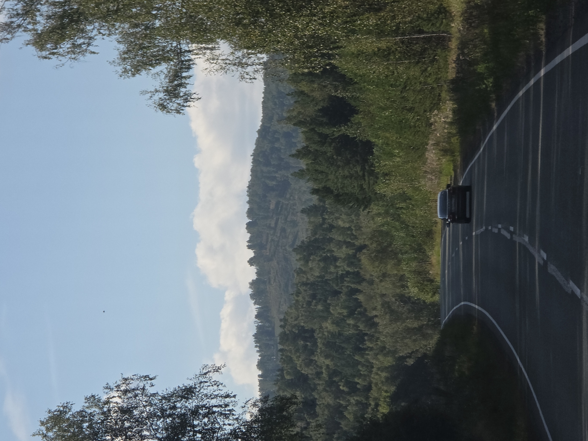 The image shows a dark-colored car driving on a winding asphalt road through a forested hilly landscape. The road has white lane markings. On the left side of the road, a grassy slope is covered with shadows. On the right side, there is a dense forest with deciduous trees and some evergreens. In the background, there are more hills covered with forests, and the sky is blue with some white clouds. The image appears to be taken in Novoandreevka, Russia, during daylight hours, likely in the summer or early autumn given the greenery. There are no people visible in the image.